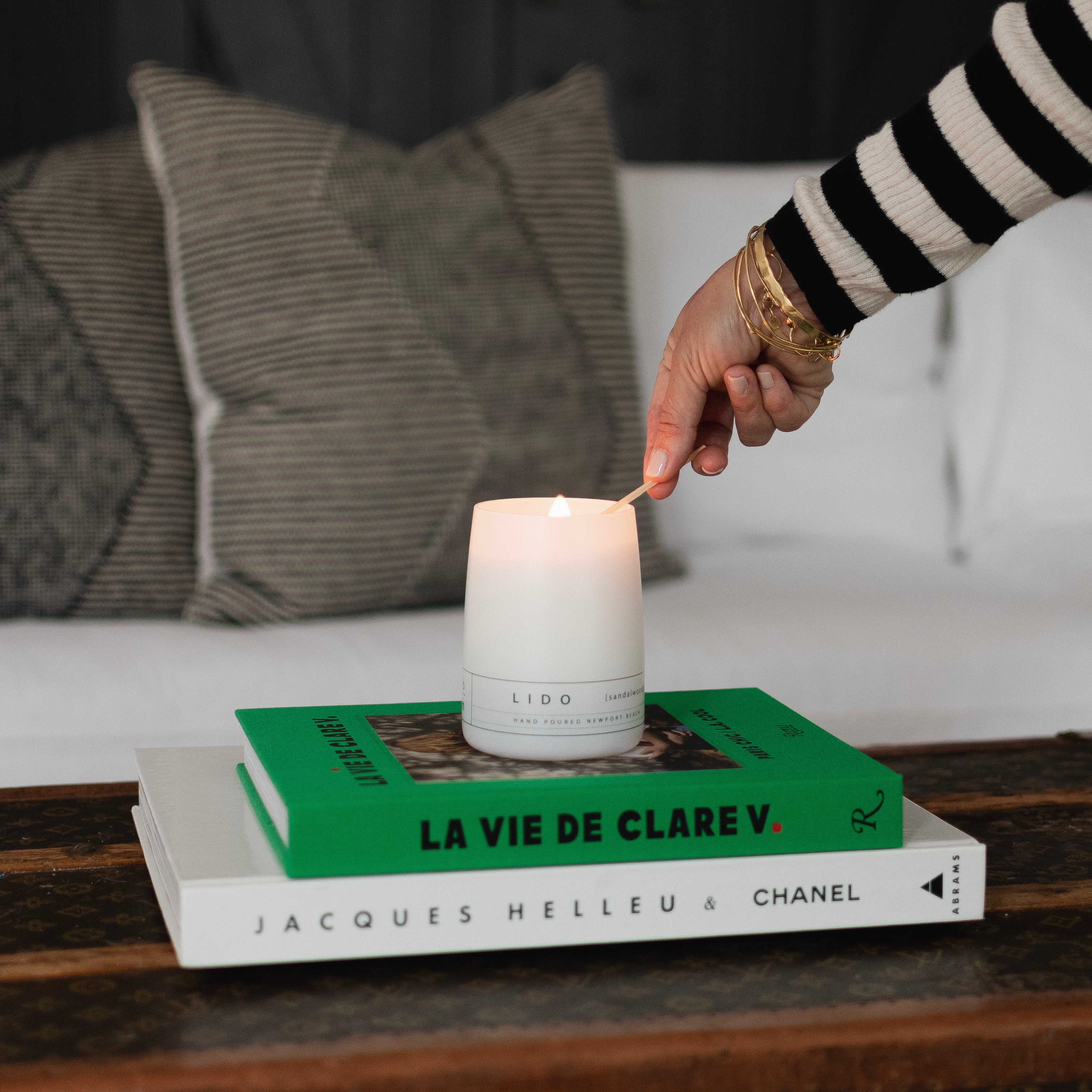 A white LIDO scented candle being lit with a match, sitting atop stacked fashion books including La Vie de Clare V. and Jacques Helleu & Chanel on a vintage trunk Newport Beach