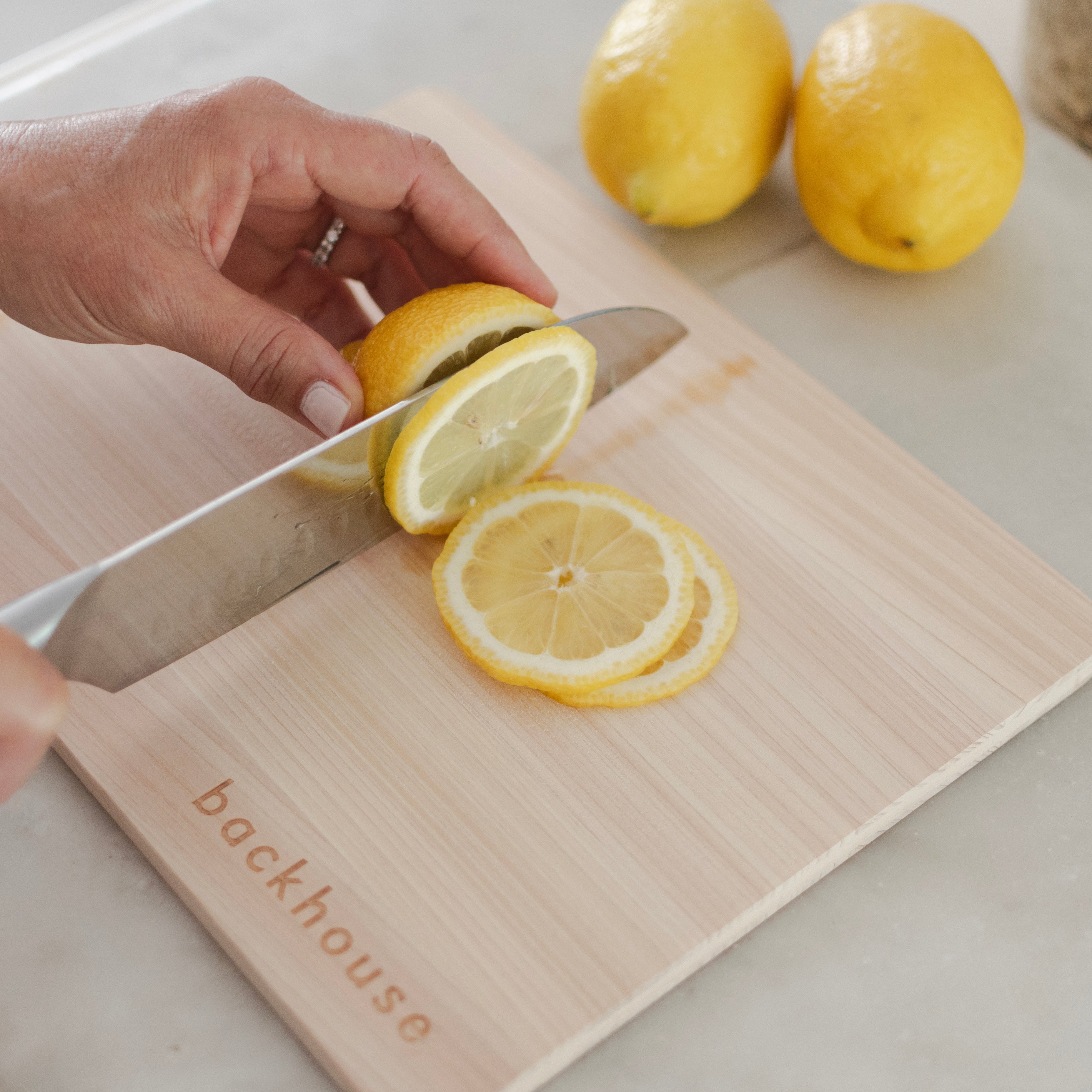 Hands slicing fresh yellow lemons on a Backhouse branded maple cutting board on a white marble kitchen counter with whole lemons in the background.