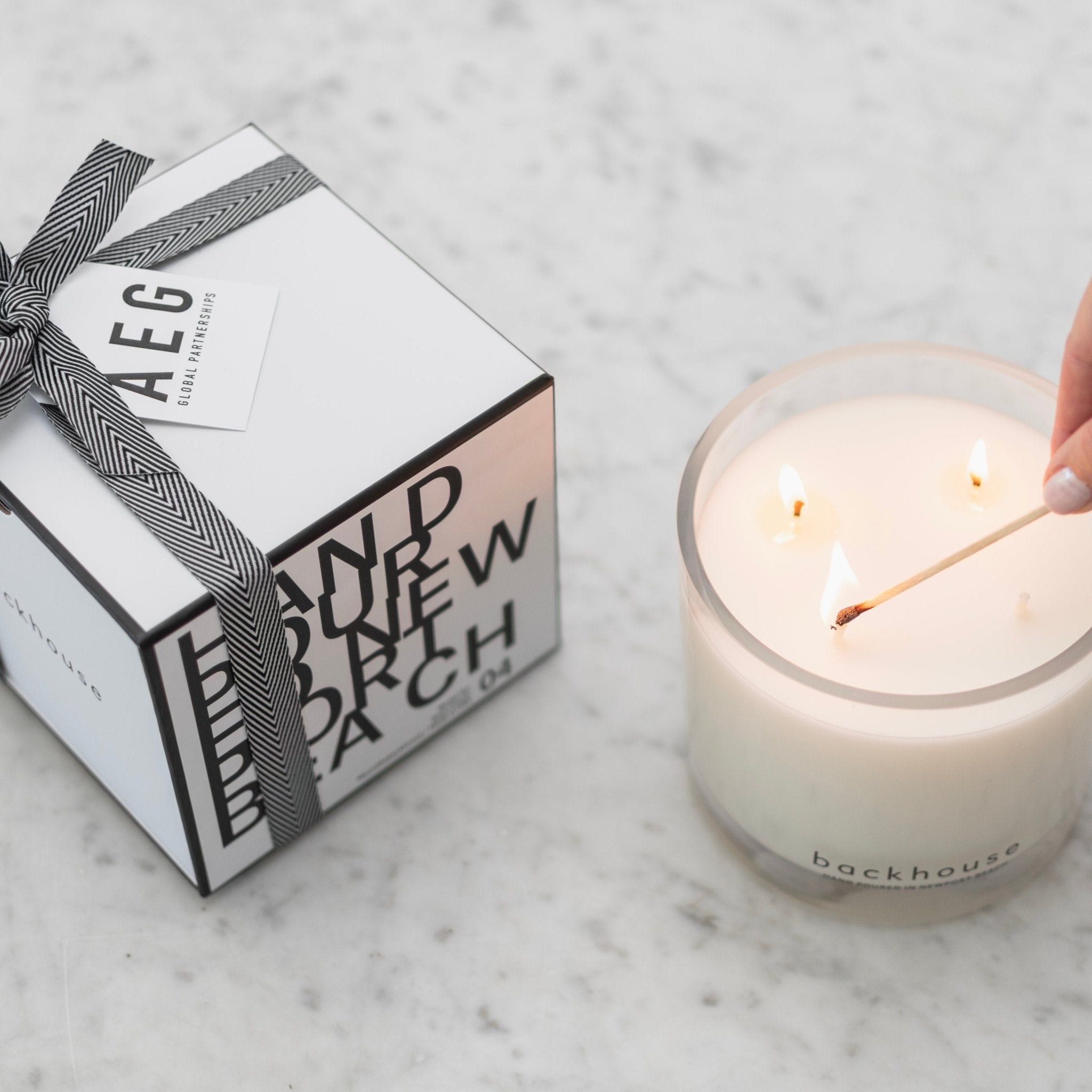 Backhouse Fragrances hurricane candle being lit with a match beside an AEG Global Partnerships branded corporate gift box on marble