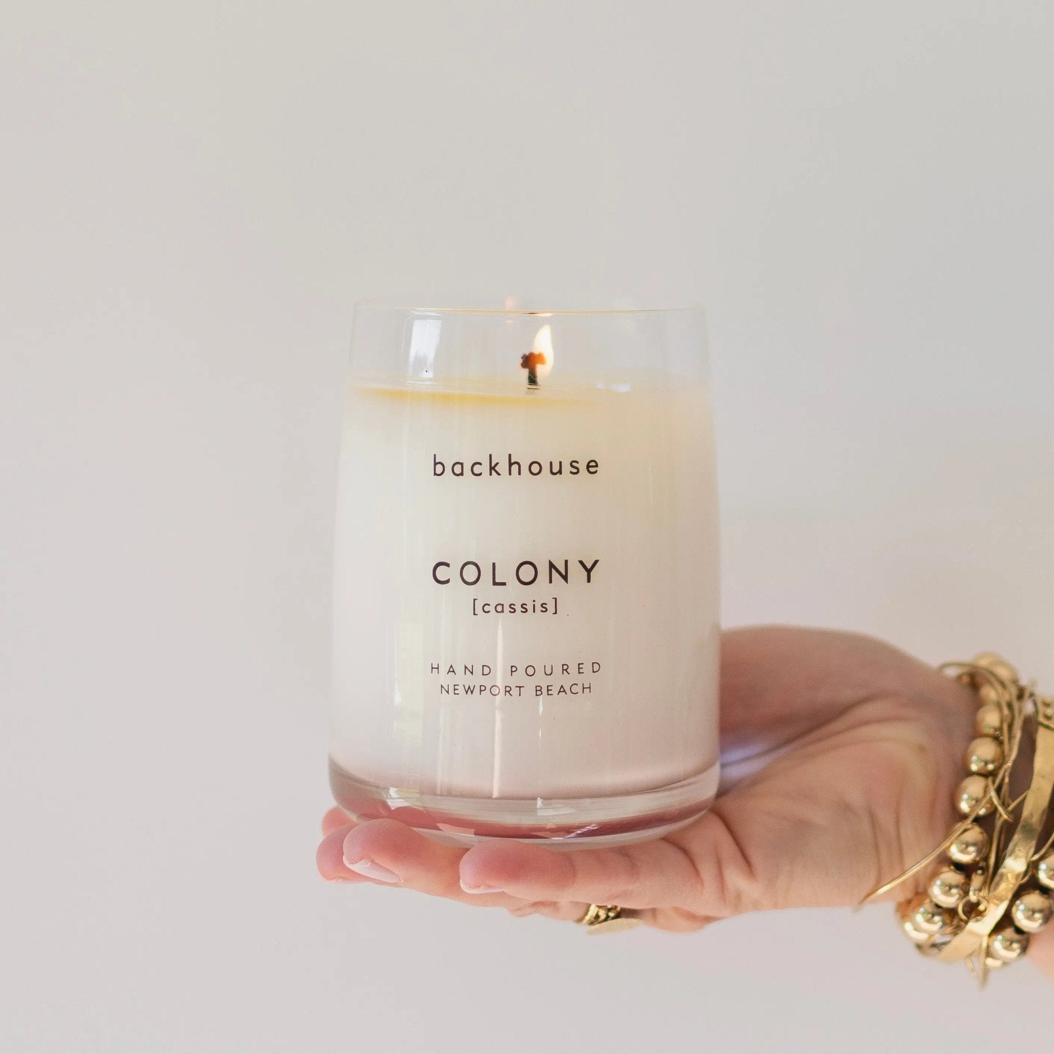 Hand holding the Colony cassis candle by backhouse while it burns against a white background in its classic glass vessel.