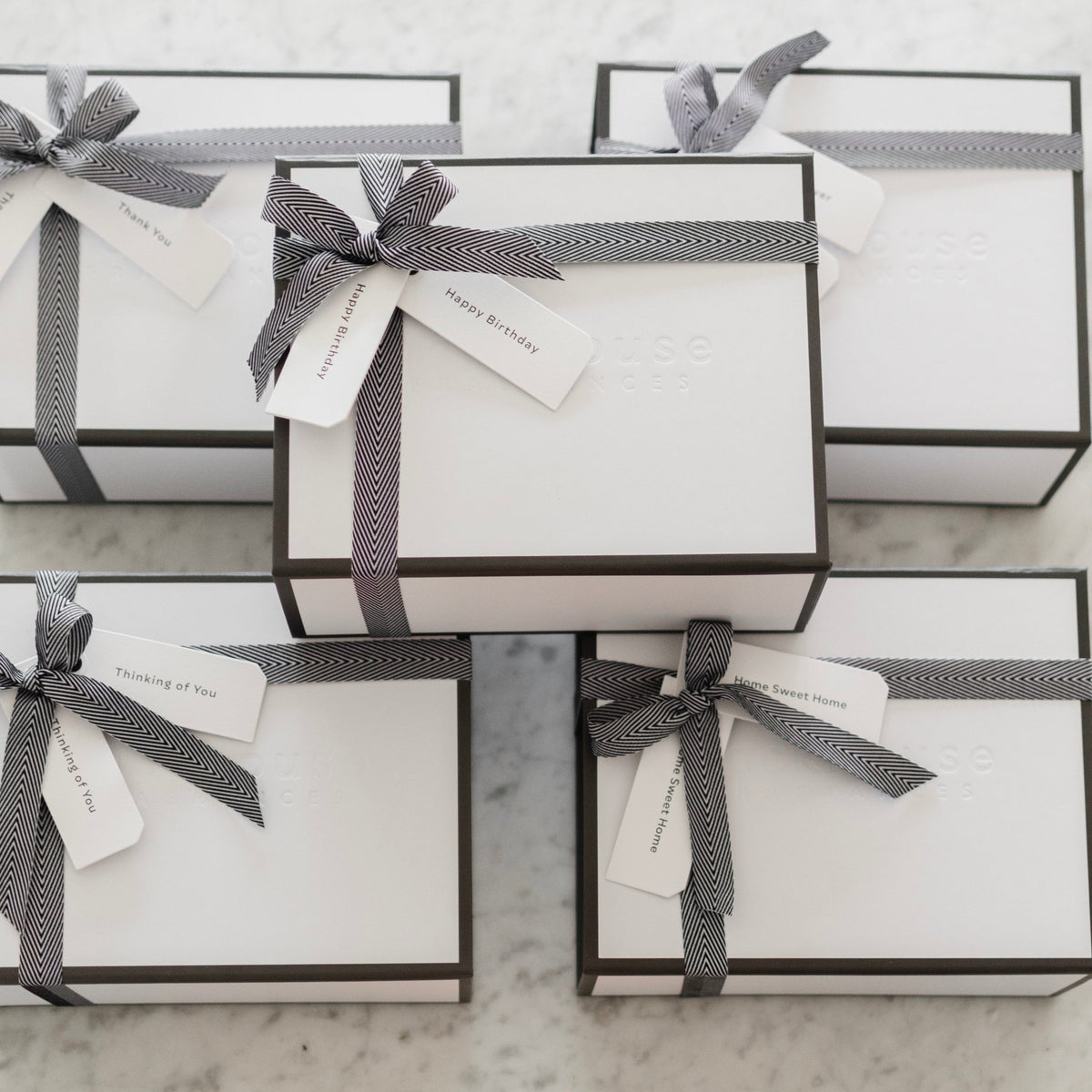 Several elegant white gift boxes with black trim and black herringbone ribbon bows, tagged with Happy Birthday, Thinking of You, Thank You, and Home Sweet Home labels, arranged on a marble surface with a small candle nearby.