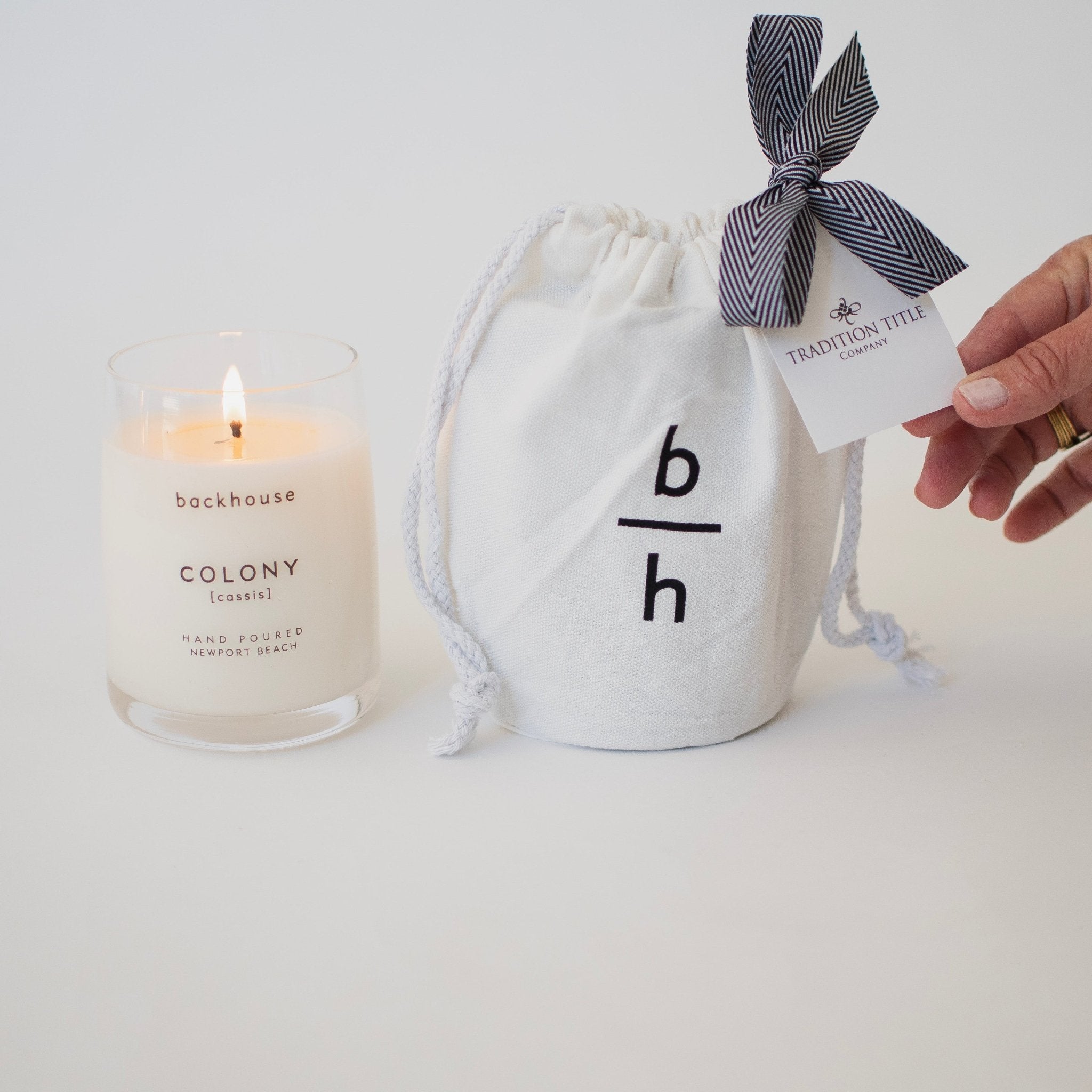Backhouse Colony cassis candle lit beside a branded canvas bag with Tradition Title Company corporate gift tag and herringbone ribbon