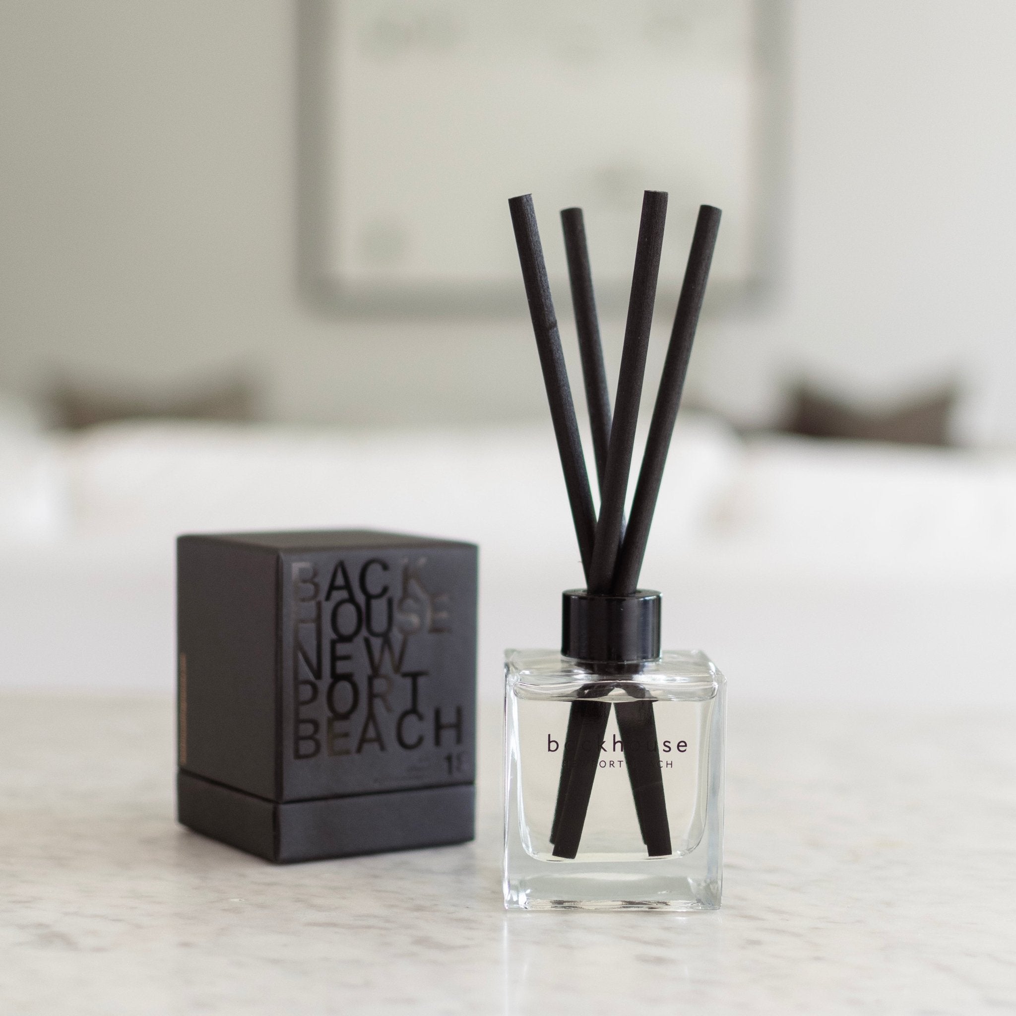 Backhouse Newport Beach LIDO reed diffuser, sandalwood scent, in a clear glass bottle with black reeds and gift box on a marble countertop.
