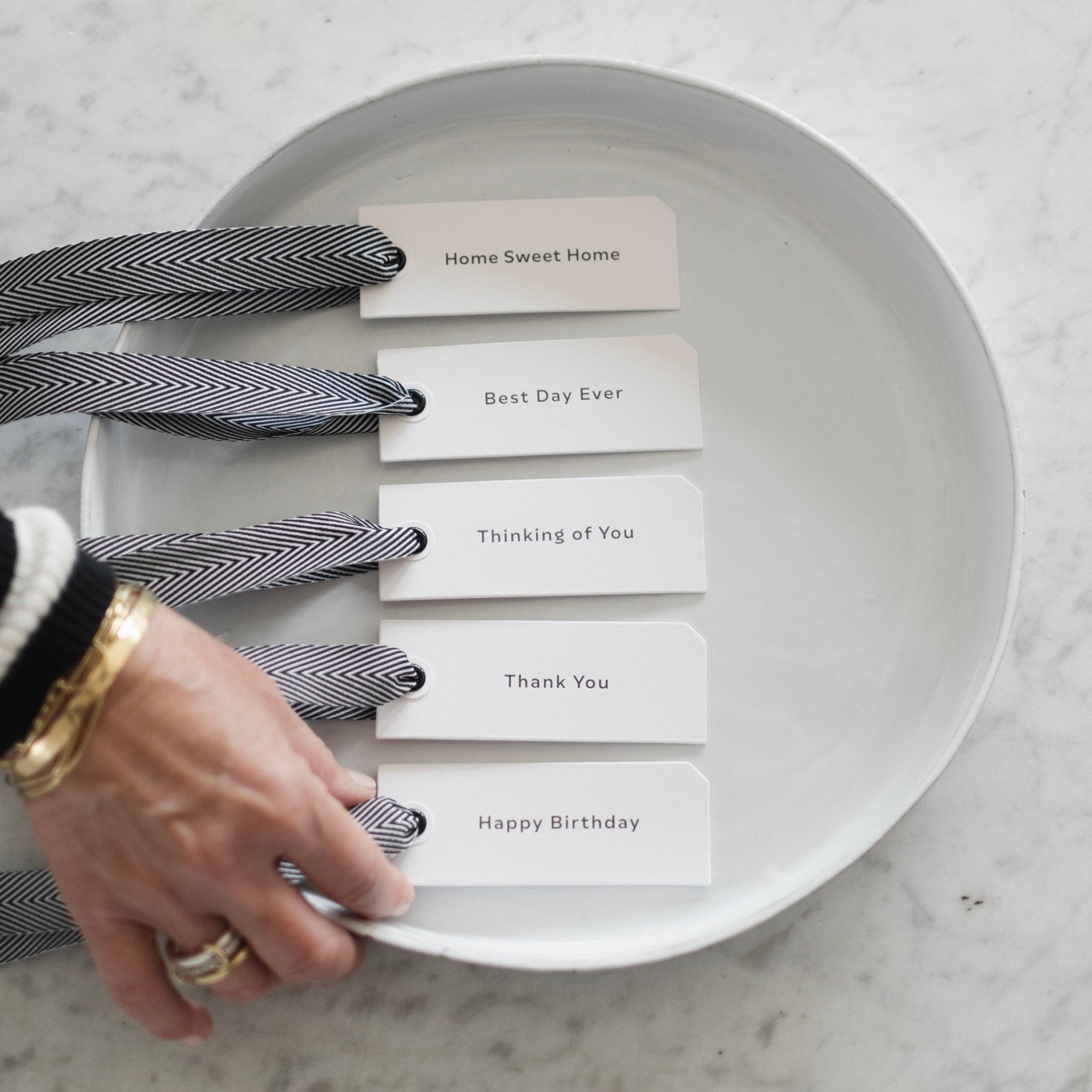 Five minimalist white gift tags with black herringbone ribbon reading Home Sweet Home, Best Day Ever, Thinking of You, Thank You, and Happy Birthday arranged on a white round tray on a marble surface, with a womans hand placing the bottom tag.