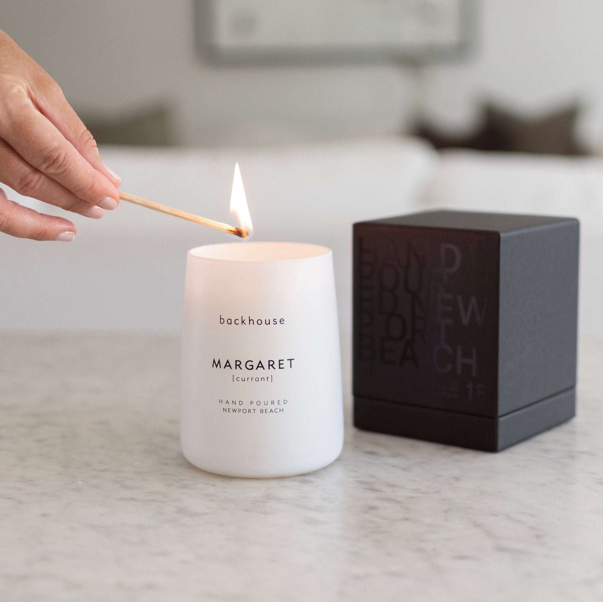 Hand lighting Backhouse Fragrances Margaret currant scented candle in matte white vessel next to matching black gift box, hand-poured luxury candle Newport Beach