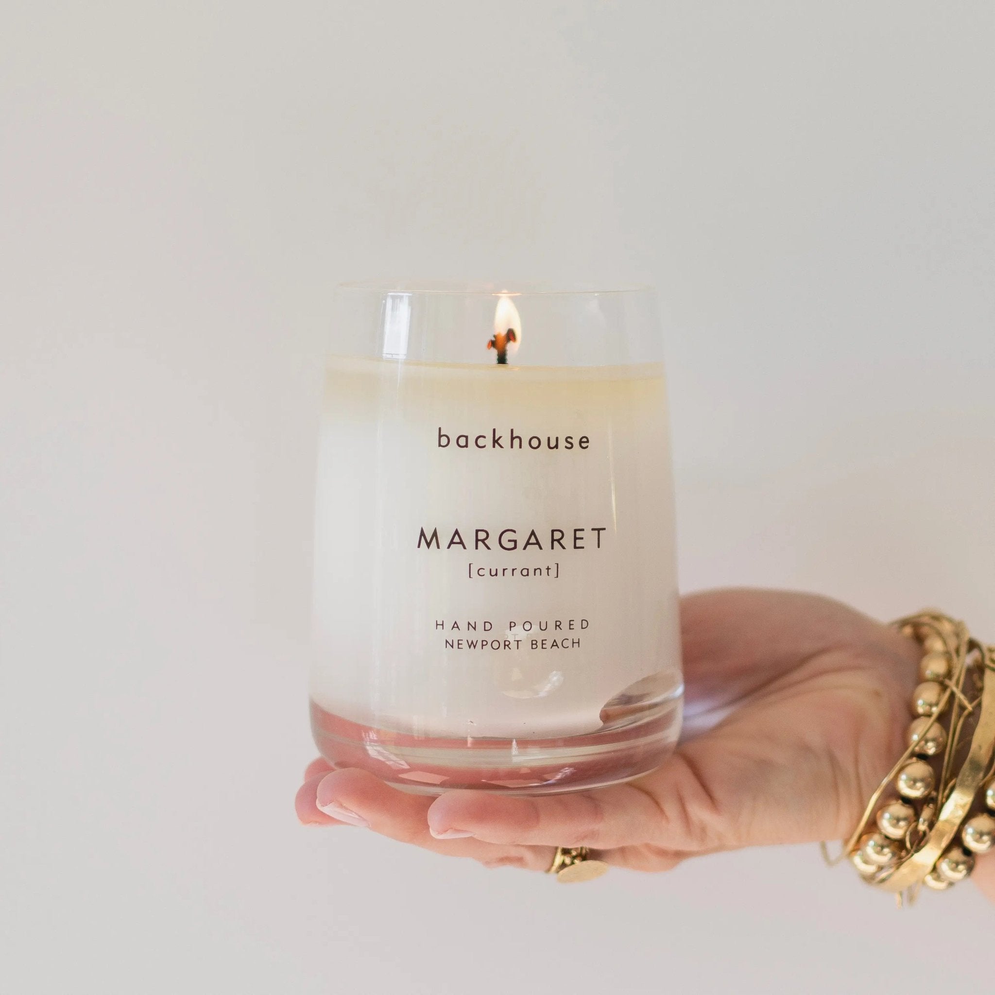 Hand holding the Margaret currant candle by backhouse while it burns against a white background in its classic glass vessel.