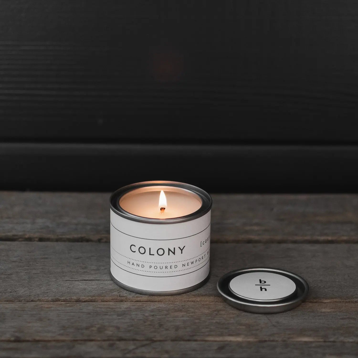 Mini Colony cassis candle by backhouse burning on a wood surface with the metal lid placed beside it against a dark background.