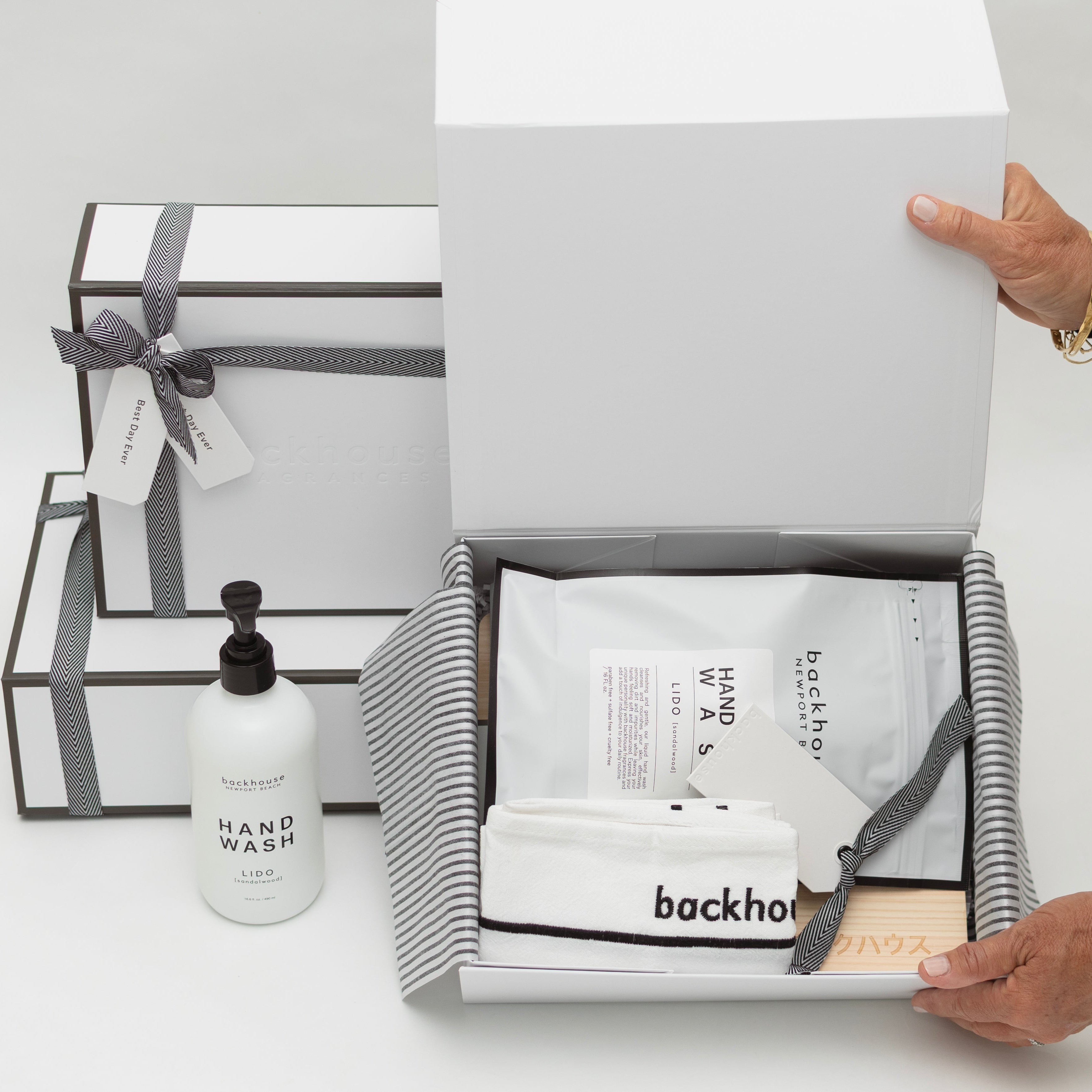 A woman in a black and white striped sweater opening a white gift box containing Backhouse branded hand wash, towels, and bath products, with additional wrapped gift boxes stacked nearby on a white surface.