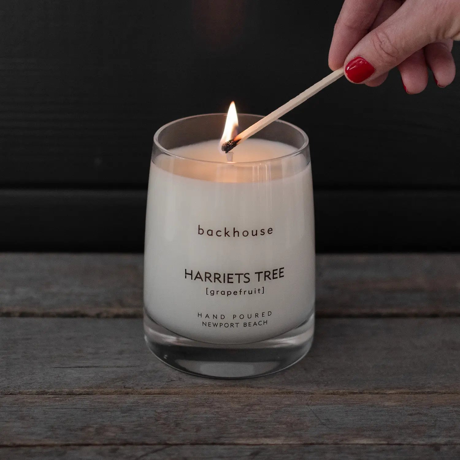 Hand lighting the Harriets Tree grapefruit hurricane candle by backhouse with a match against a dark background.