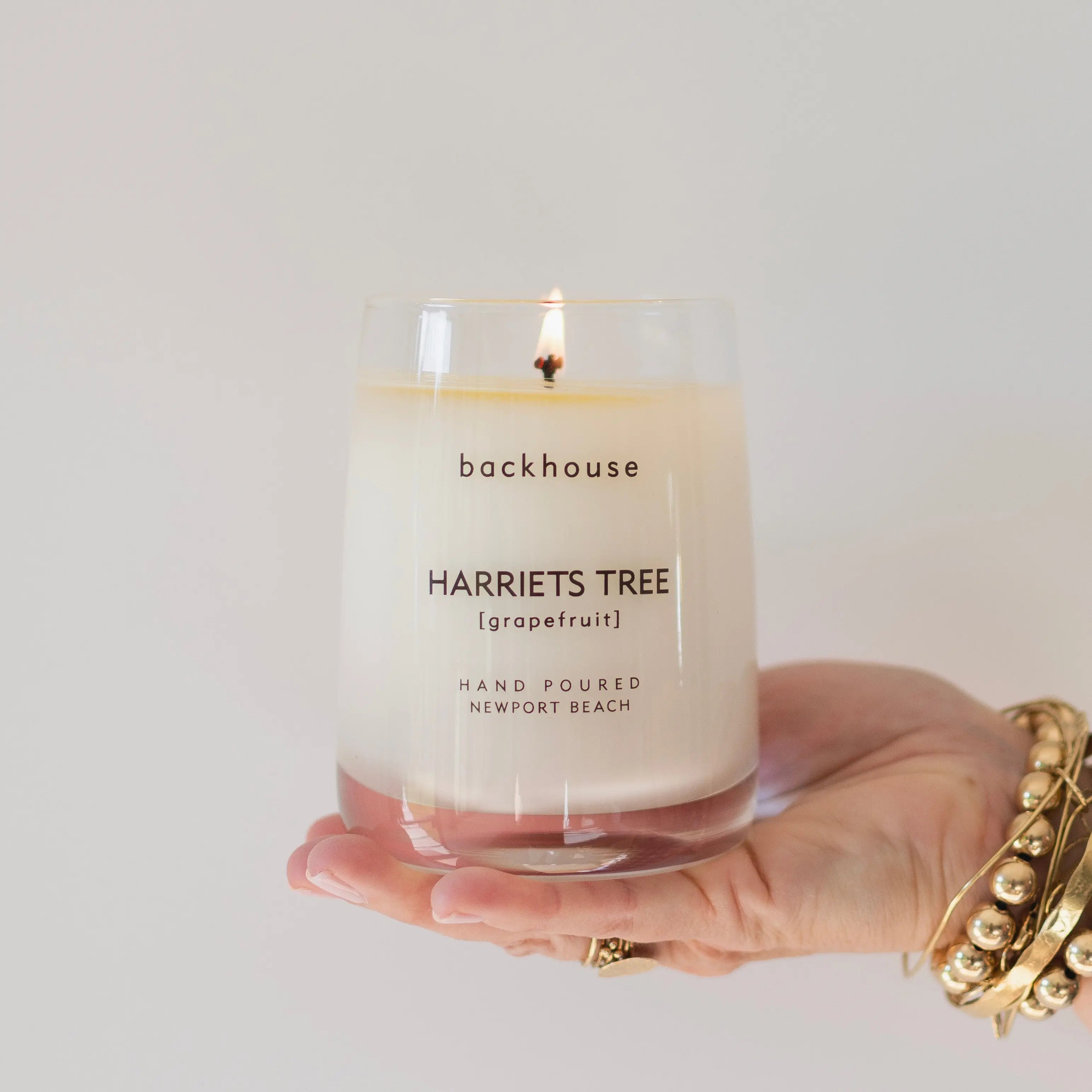 Hand holding the Harriets Tree grapefruit hurricane candle by backhouse while it burns against a white background in its classic glass vessel.