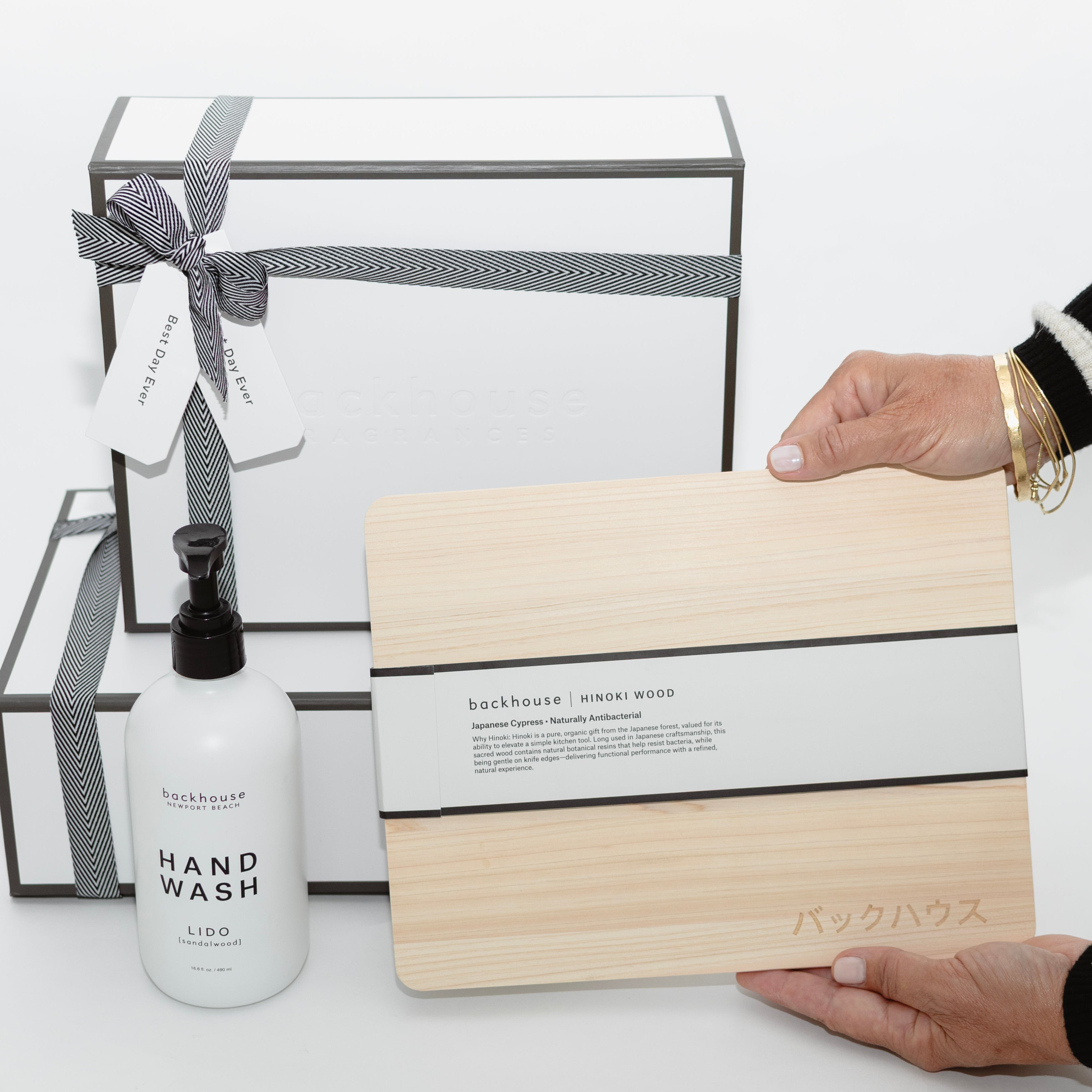 A woman in a black and white striped sweater holding a backhouse hinoki wood cutting board next to a backhouse hand wash soap bottle and two white gift boxes tied with black and white ribbon on a white surface.