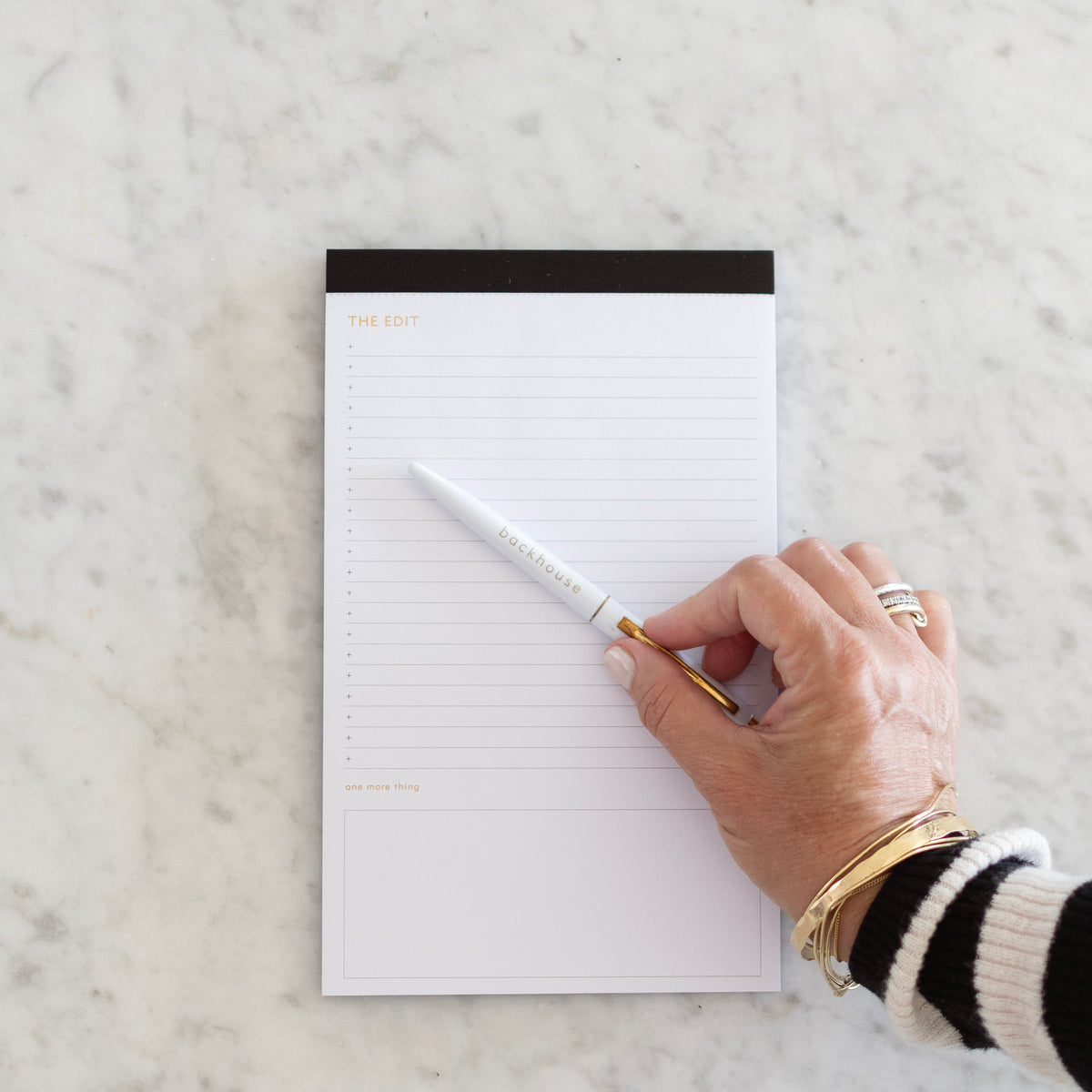 White "The Edit" notepad by backhouse with gold lettering and a black top binding, shown on a marble surface with a hand holding a white and gold backhouse pen.