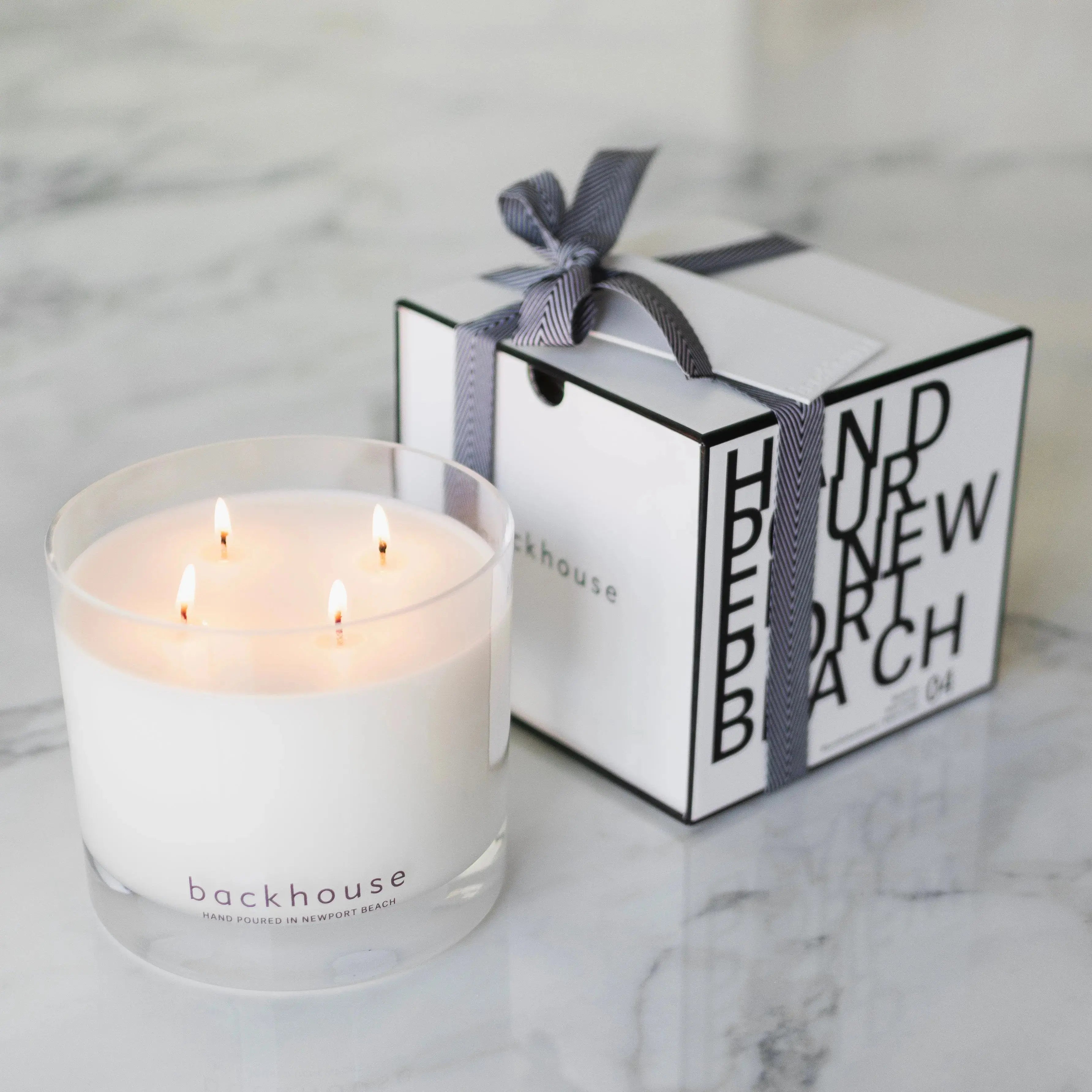The Big BH sandalwood 4-wick candle by backhouse burning on a white surface beside its cylinder box wrapped with ribbon.