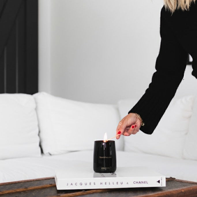 Person lighting a candle on a coffee table with a book underneath.  Black and gold candle in a sandalwood scent.  Modern and timeless vessel.