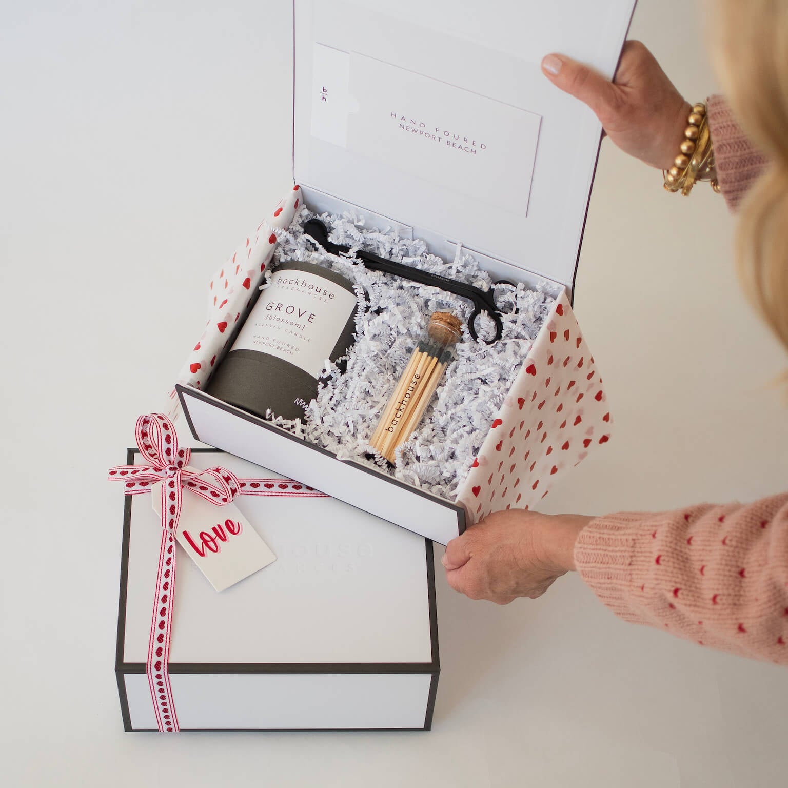Person opening a gift box with candle + wick trimmer + matches inside on a light background