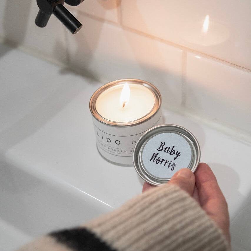 Candle in a tin labeled 'Baby Norris' held by a hand over a sink.