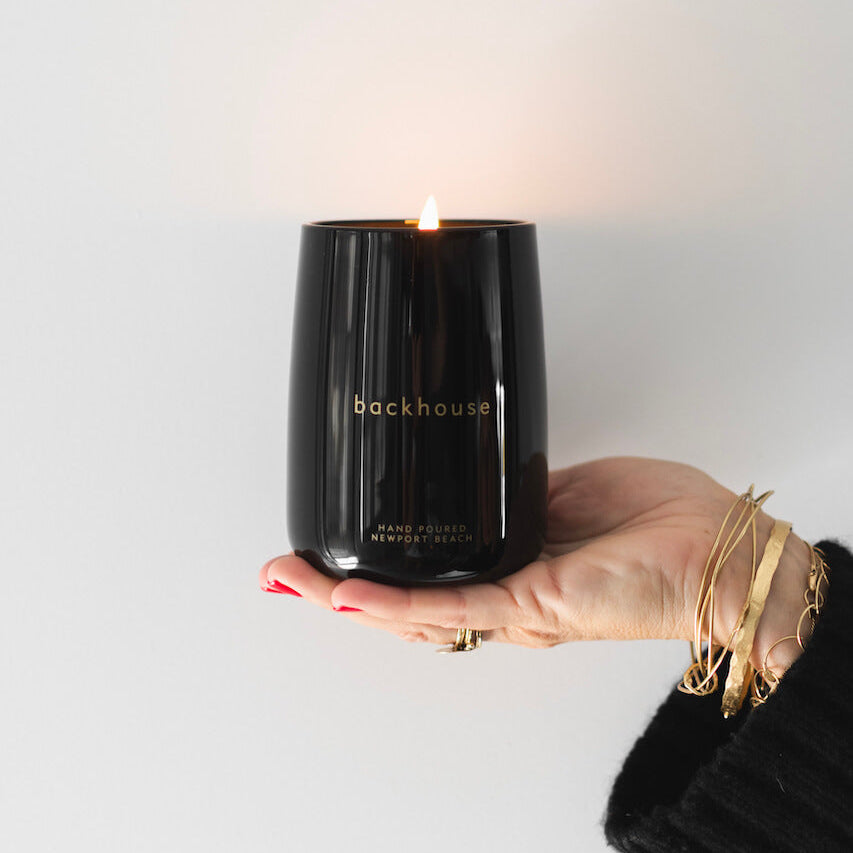 Hand holding the Black + Gold pine candle by backhouse while it burns against a white background in a black glass vessel.