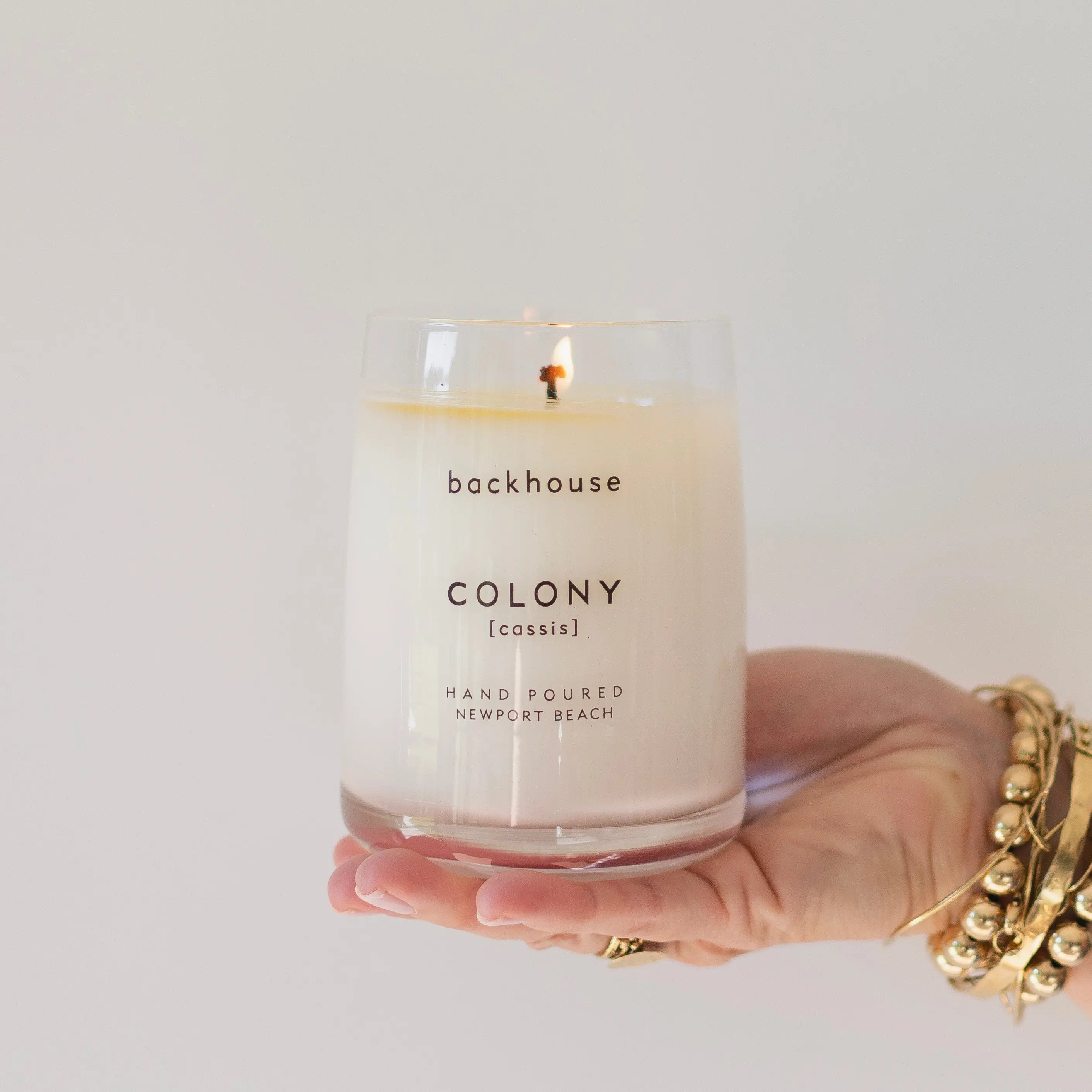 Hand holding the Colony cassis candle by backhouse while it burns against a white background in its classic glass vessel.