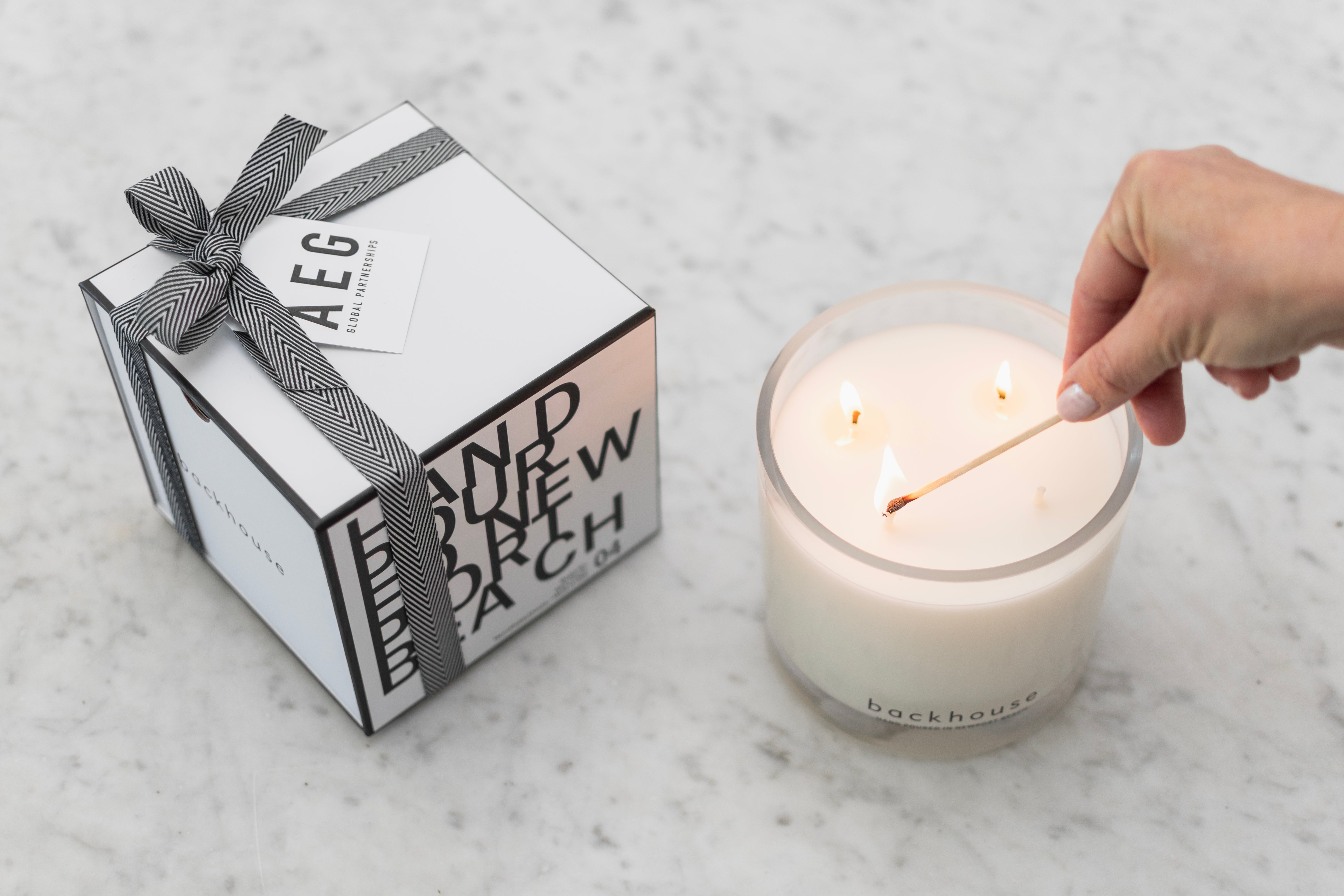 Backhouse Fragrances three-wick candle hand-poured in Newport Beach, being lit beside a custom AEG-branded gift box.