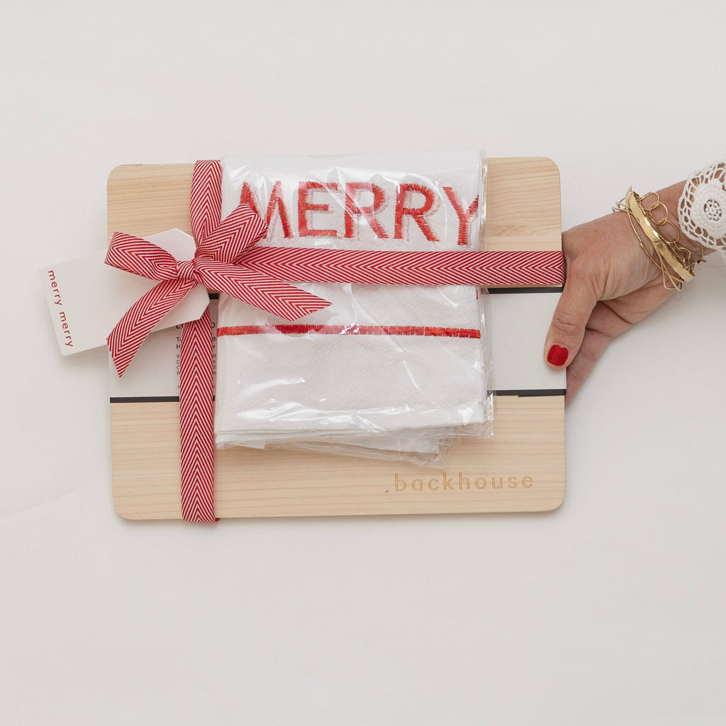 A wooden cutting board gift set from Backhouse featuring a white embroidered Merry tea towel tied with a red and white herringbone ribbon bow, held by a woman's hand with red nail polish and gold bracelets.
