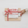 A wooden Backhouse branded cutting board gift set wrapped with a red and white herringbone ribbon bow and a Merry embroidered towel, held by a woman's hand with red nails and gold bracelets on a white surface.