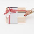 A wooden Backhouse desk organizer gift set wrapped with a red herringbone ribbon and bow, featuring a notepad and pen, held by a woman's hand with red nails and gold bracelets, against a white background.