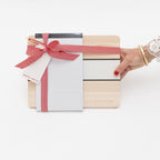 A wooden Backhouse desk organizer gift set wrapped with a red herringbone ribbon and bow, featuring a notepad and pen, held by a woman's hand with red nails and gold bracelets, against a white background.