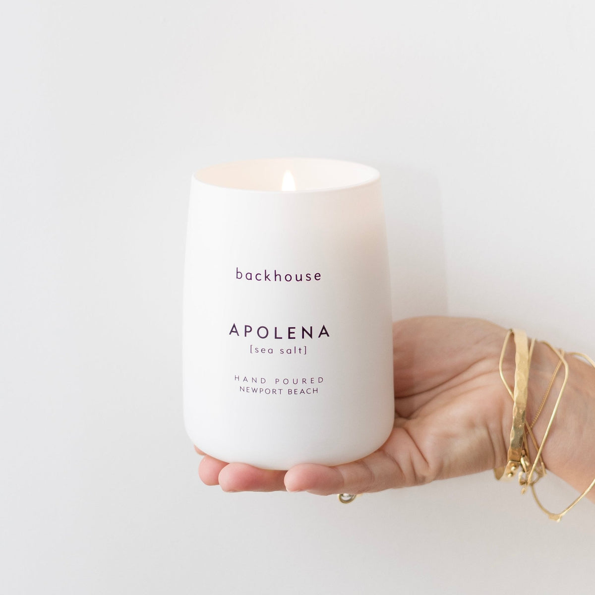 A hand wearing gold bracelets holds a white matte ceramic Backhouse Apolena sea salt scented hand poured candle against a clean white background.