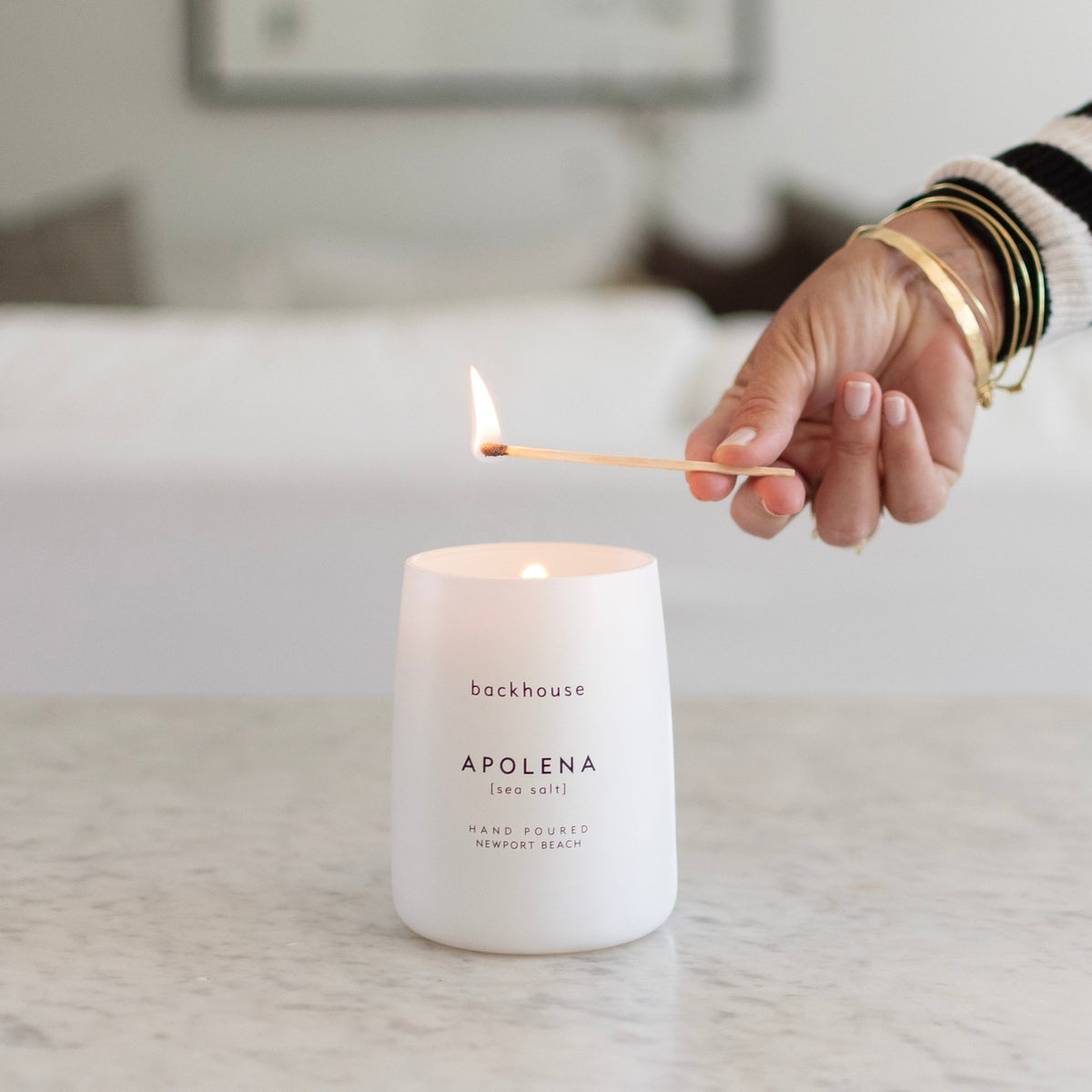 A hand wearing gold bracelets and a black and white striped sweater lighting a backhouse Apolena sea salt hand poured candle in a white matte vessel on a marble surface with a white sofa in the background.