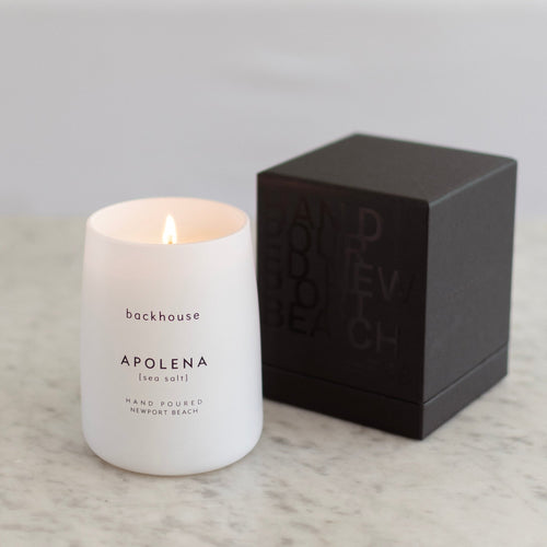 A lit white matte glass Backhouse Apolena sea salt scented candle hand poured in Newport Beach sitting on a marble surface next to its dark charcoal gift box.