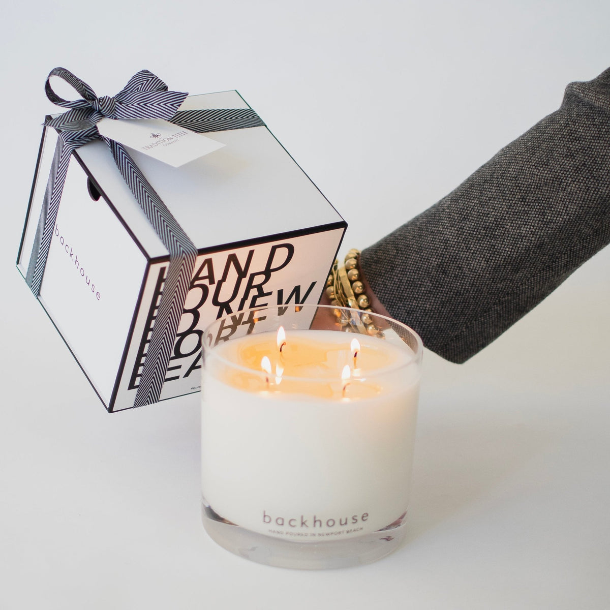 Backhouse Fragrances large hurricane candle with three lit wicks beside a closed gift box with herringbone ribbon