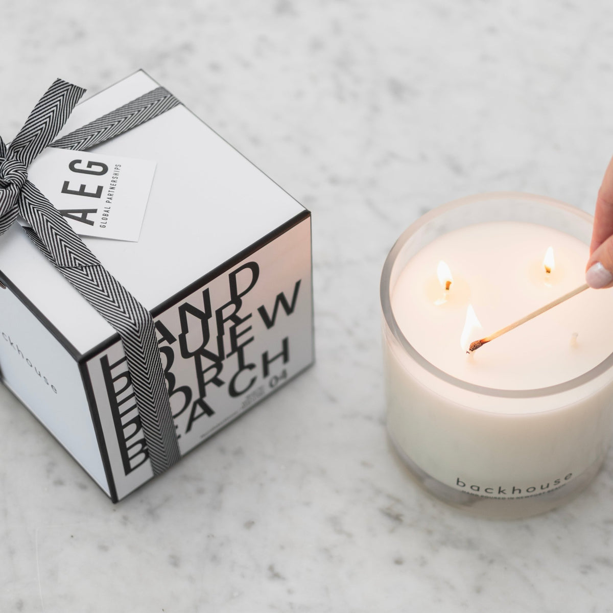 Backhouse Fragrances hurricane candle being lit with a match beside an AEG Global Partnerships branded corporate gift box on marble