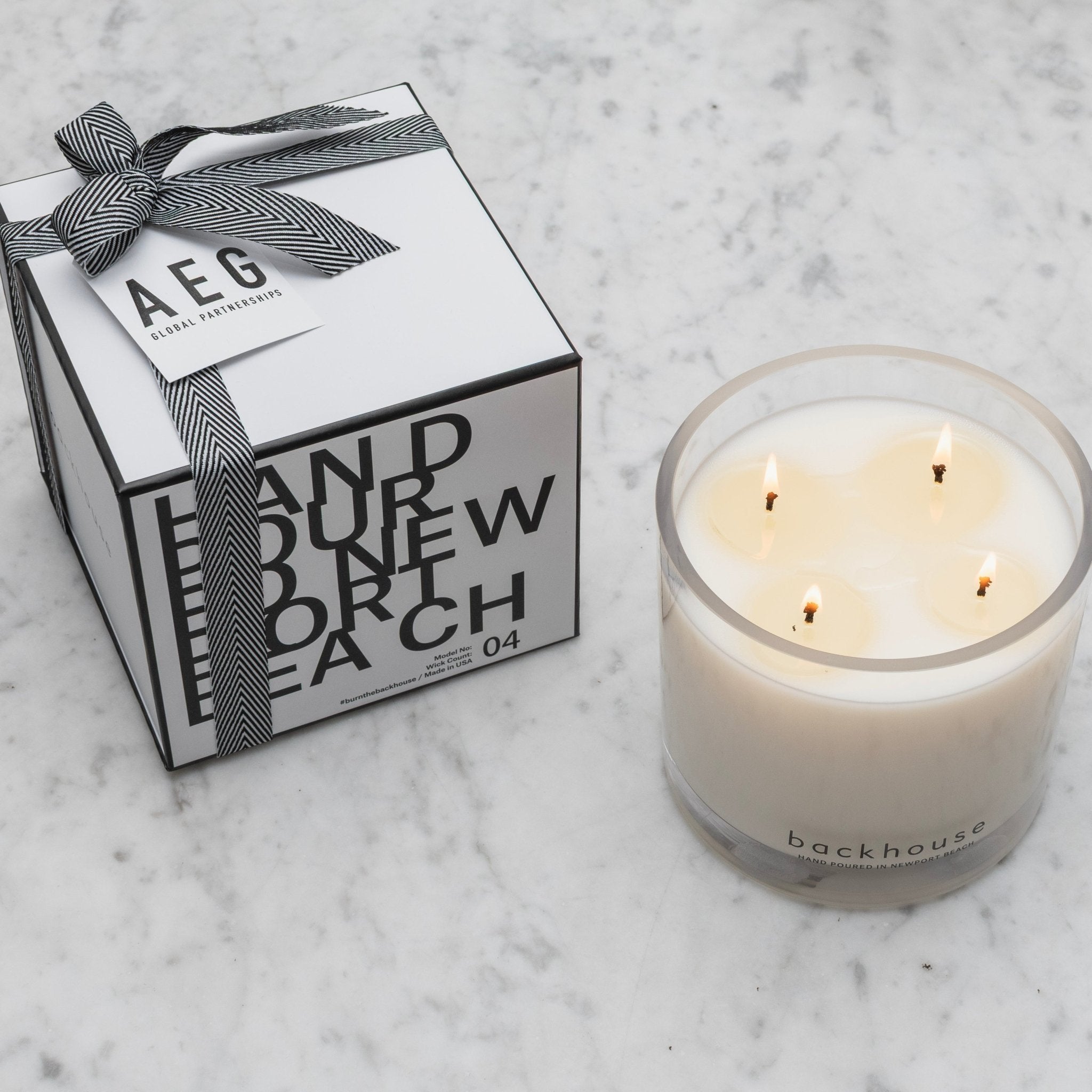 Backhouse Fragrances lit hurricane candle beside an AEG Global Partnerships branded corporate gift box with herringbone ribbon on marble
