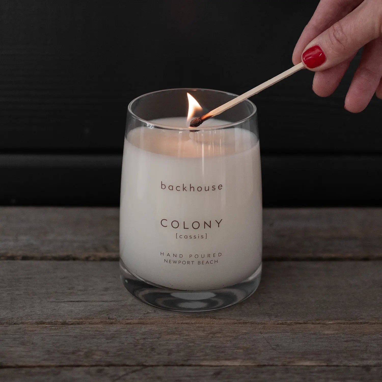 Hand lighting the Colony cassis candle by backhouse with a match against a dark background.