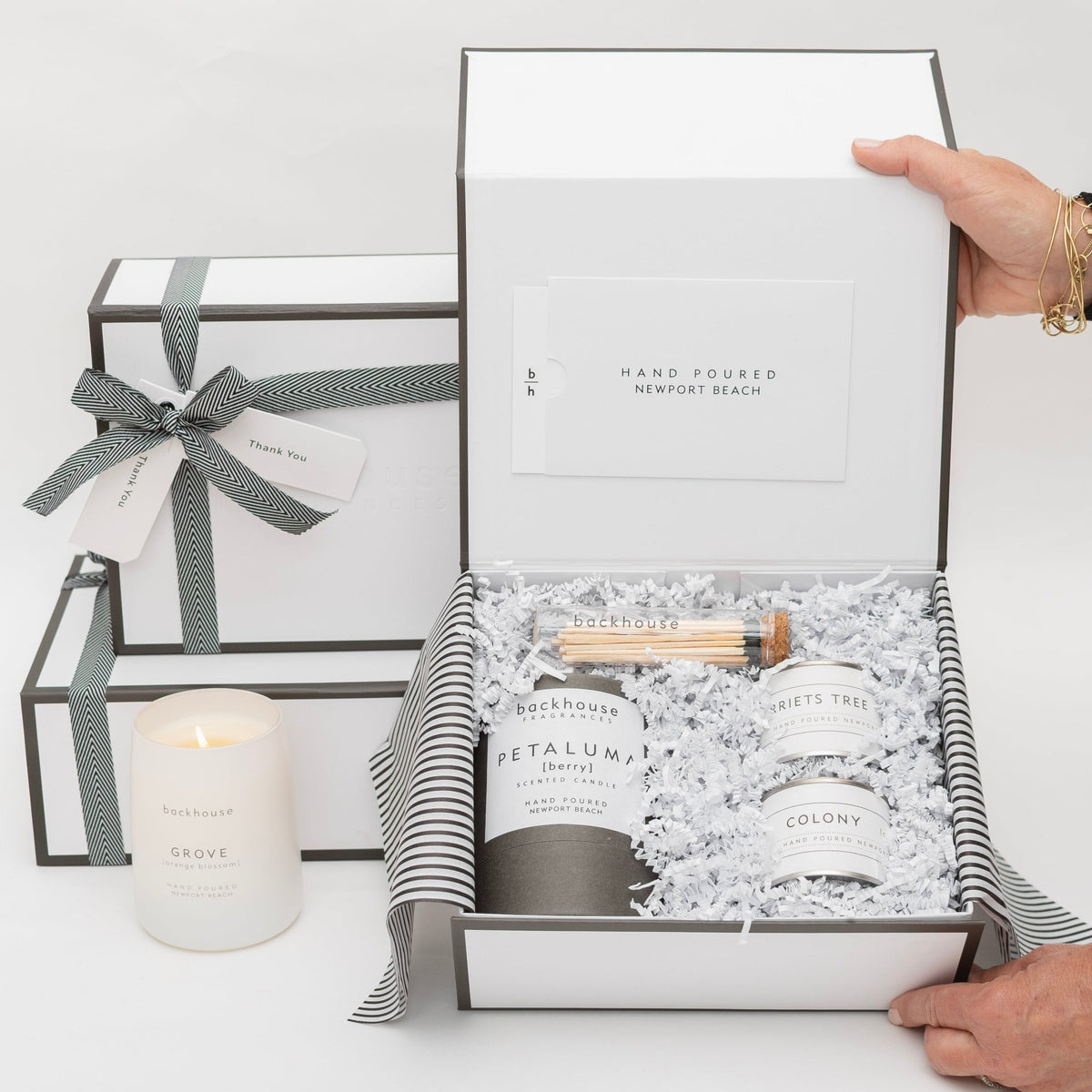 A person opening a elegant white gift box containing Backhouse Fragrances candles including Petaluma berry scent alongside matches and smaller candles, with additional wrapped gift boxes stacked nearby and a lit Grove candle displayed in front.