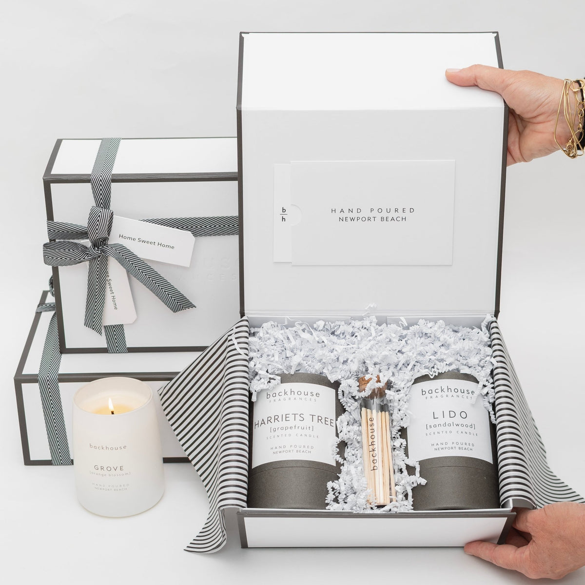 A pair of hands opening a white gift box containing Backhouse Fragrances scented candles including Harriets Tree grapefruit and Lido sandalwood scents packed in white crinkle paper beside a lit Grove candle and stacked gift boxes with black and white ribbon bows