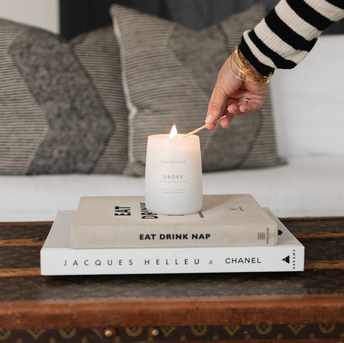 Backhouse Fragrances Grove candle with a match, the candle sitting atop two coffee table books including Jacques Helleu and Chanel and Eat Drink Nap, placed on a vintage trunk in front of a white sofa with gray pillows. Newport Beach