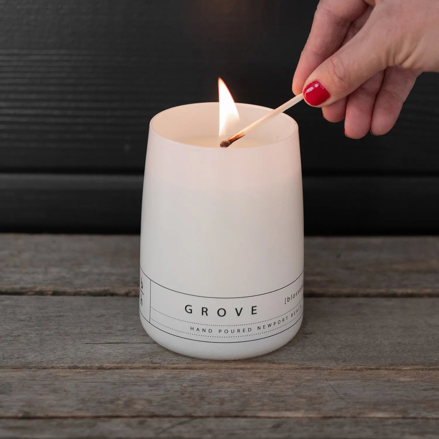 Hand lighting the Grove orange blossom candle by backhouse with a match against a dark background.