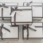Several elegant white gift boxes with black trim and black herringbone ribbon bows, tagged with Happy Birthday, Thinking of You, Thank You, and Home Sweet Home labels, arranged on a marble surface with a small candle nearby.