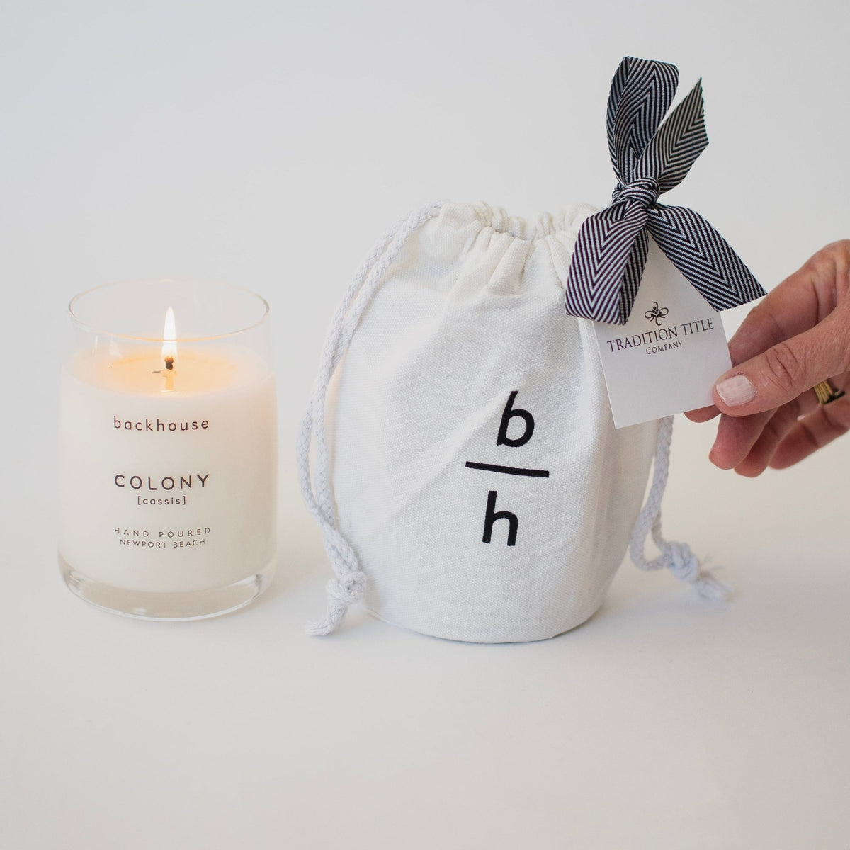 Backhouse Colony cassis candle lit beside a branded canvas bag with Tradition Title Company corporate gift tag and herringbone ribbon