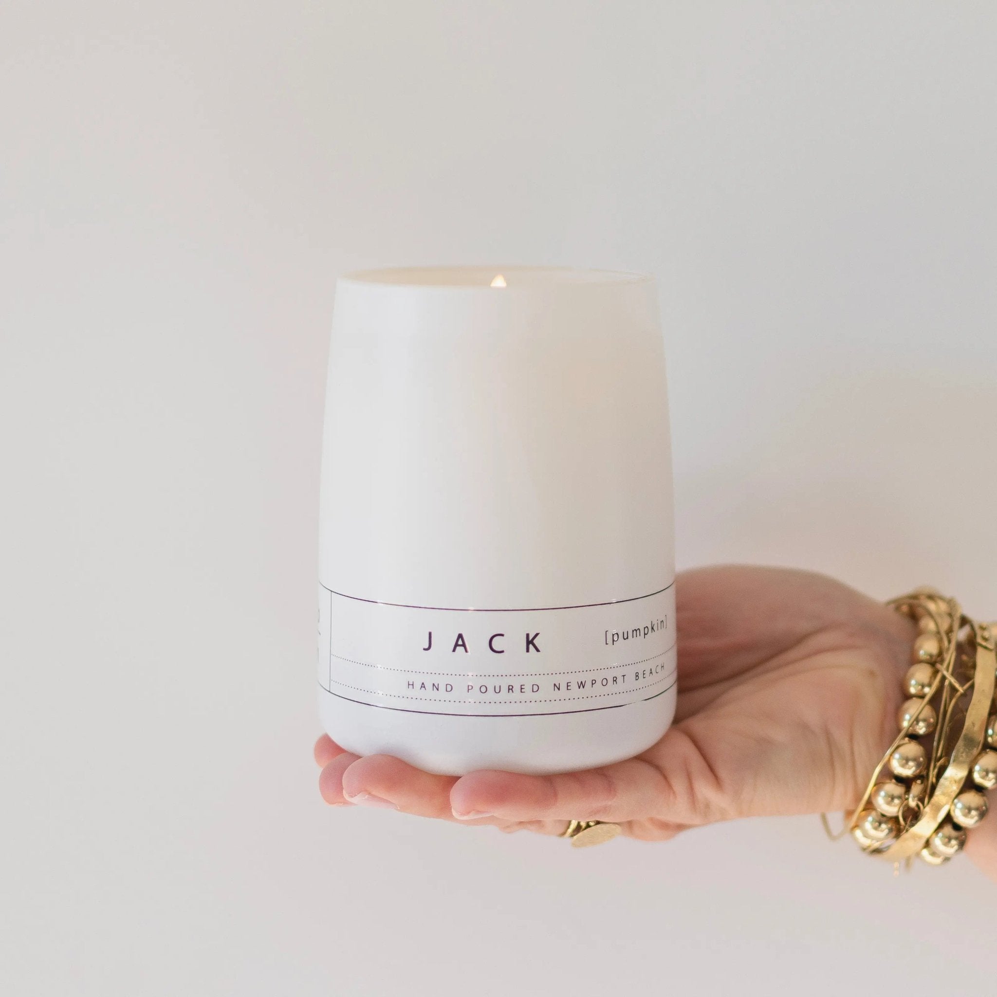 Hand holding the Jack pumpkin candle by backhouse against a white background in its classic glass vessel.