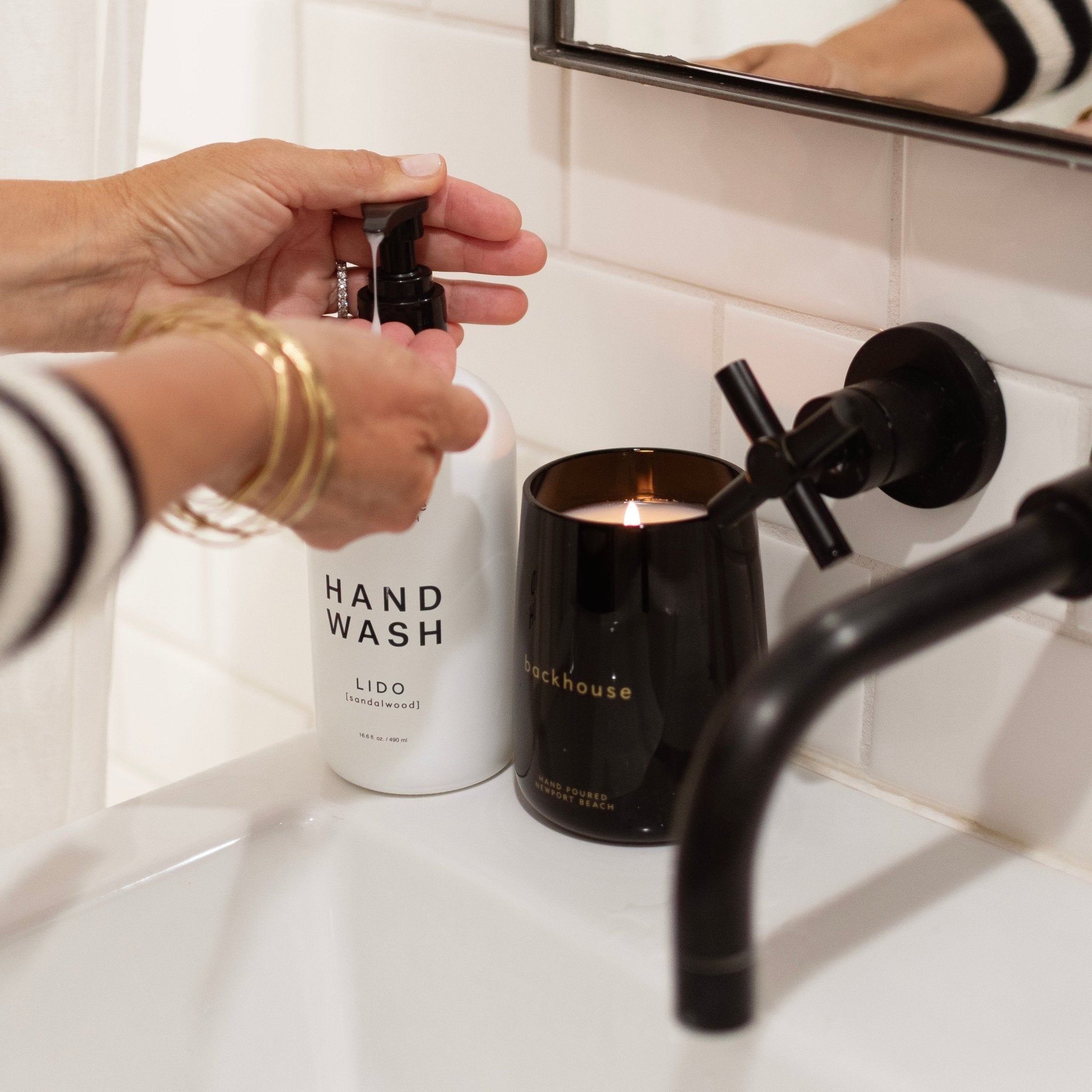 Backhouse Fragrances Lido sandalwood hand wash pump bottle next to lit black glass candle on bathroom vanity with matte black fixtures, luxury home fragrance gift set