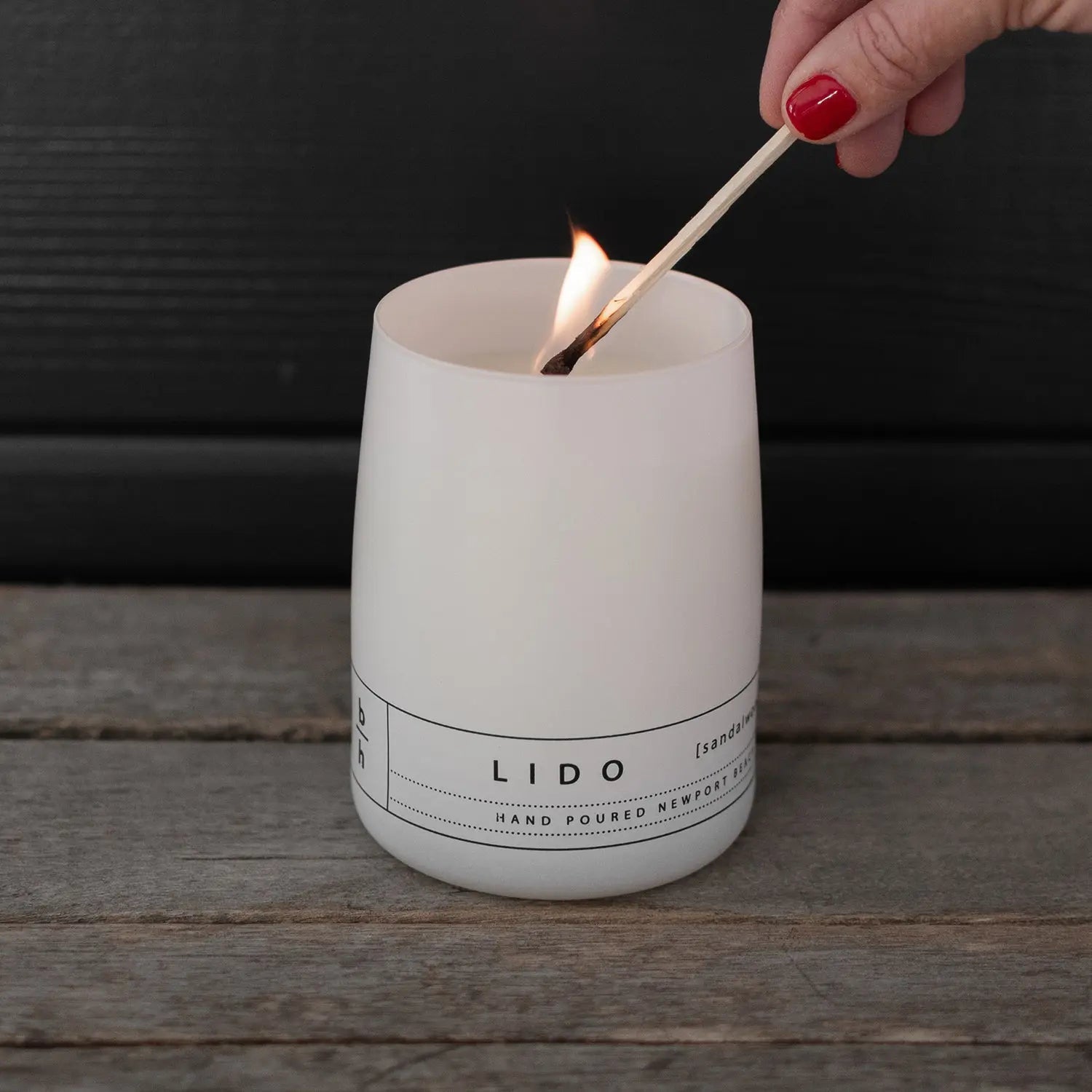 Hand lighting the Lido sandalwood hurricane candle by backhouse with a match against a dark background.