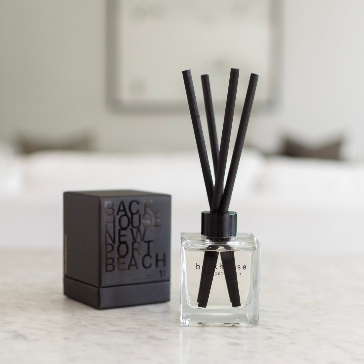Backhouse Newport Beach LIDO reed diffuser, sandalwood scent, in a clear glass bottle with black reeds and gift box on a marble countertop.