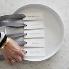 Five minimalist white gift tags with black herringbone ribbon reading Home Sweet Home, Best Day Ever, Thinking of You, Thank You, and Happy Birthday arranged on a white round tray on a marble surface, with a womans hand placing the bottom tag.