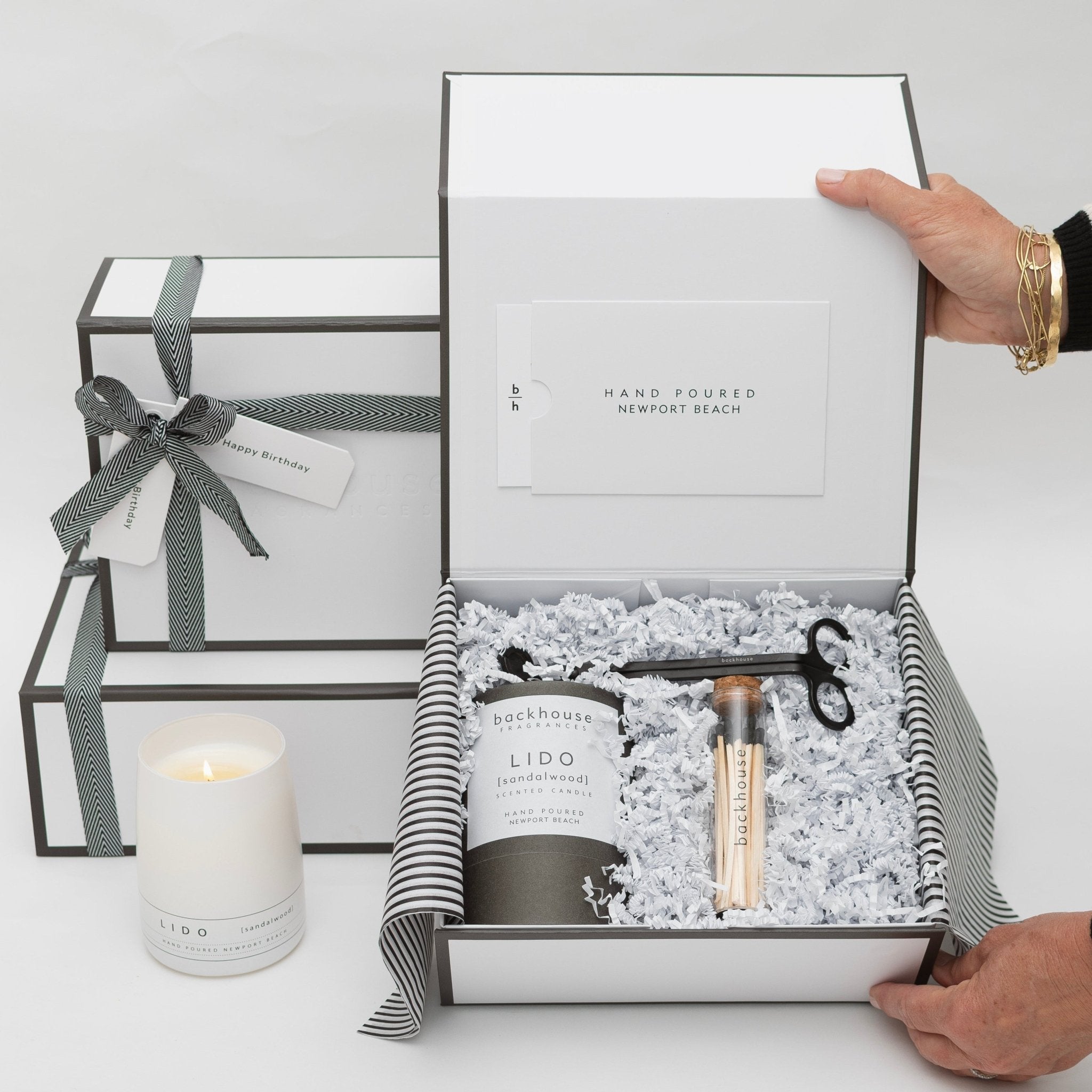 A hand poured Lido sandalwood scented candle gift set from Backhouse Fragrances displayed in an open white gift box with black and white striped tissue paper, matches, and a wick trimmer, alongside a wrapped Happy Birthday box with ribbon, on a white surface in Newport Beach.