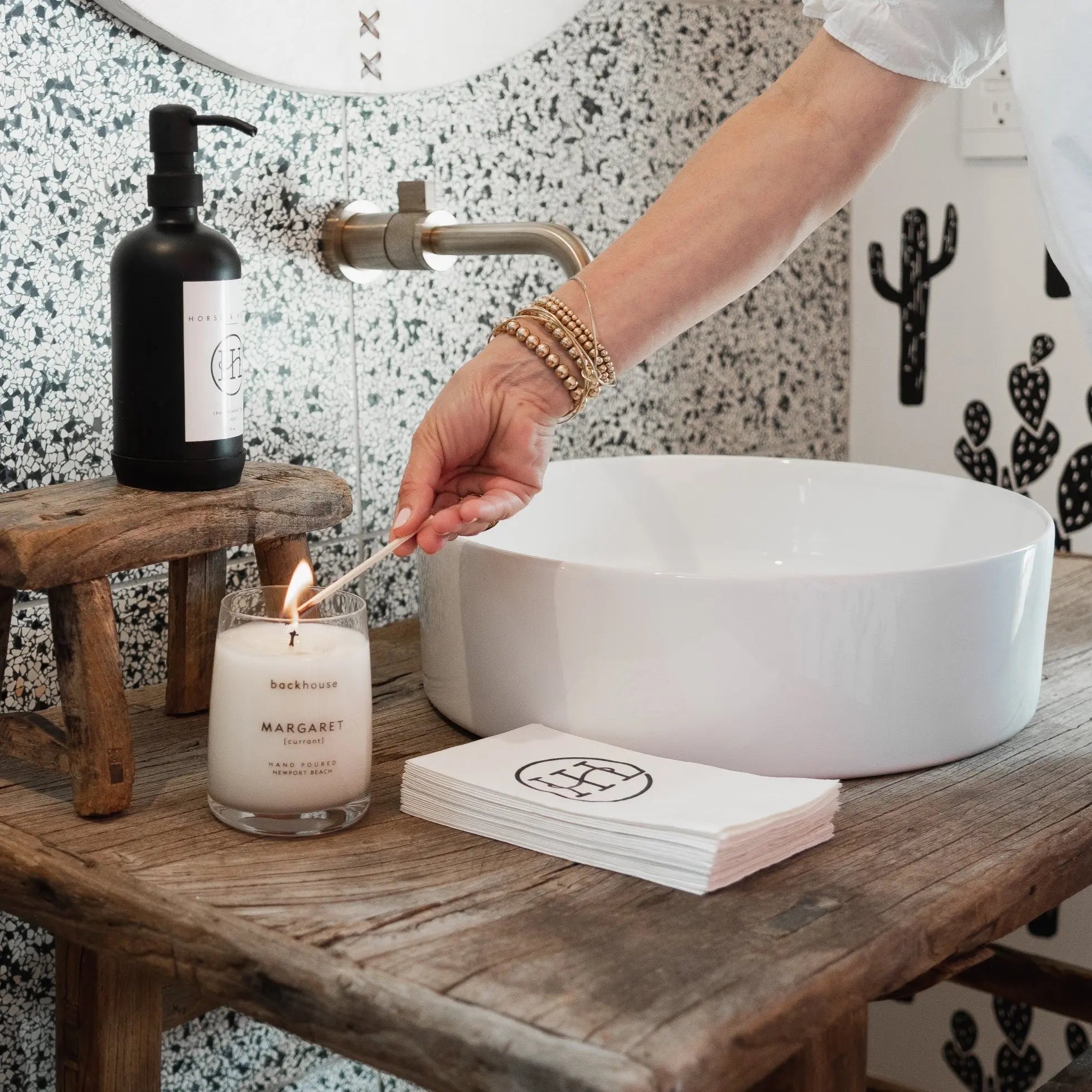 Margaret currant candle by backhouse burning on a wood bathroom counter beside a white sink.