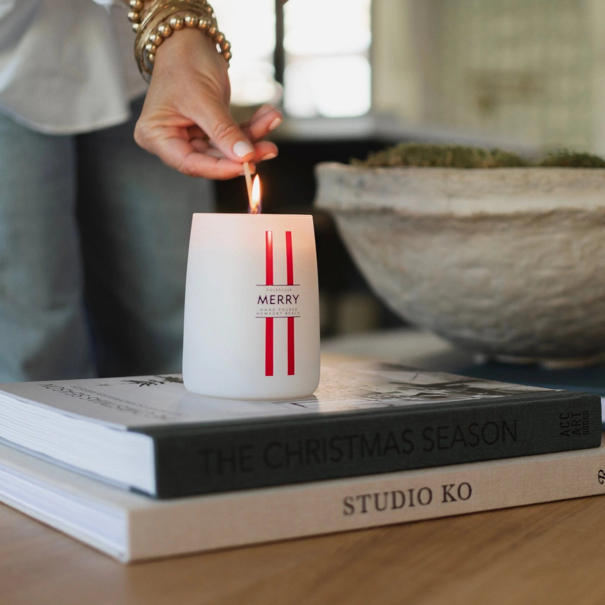 Hand lighting the Merry Mr. Douglas pine candle by backhouse while it rests on stacked coffee table books against a dark background.