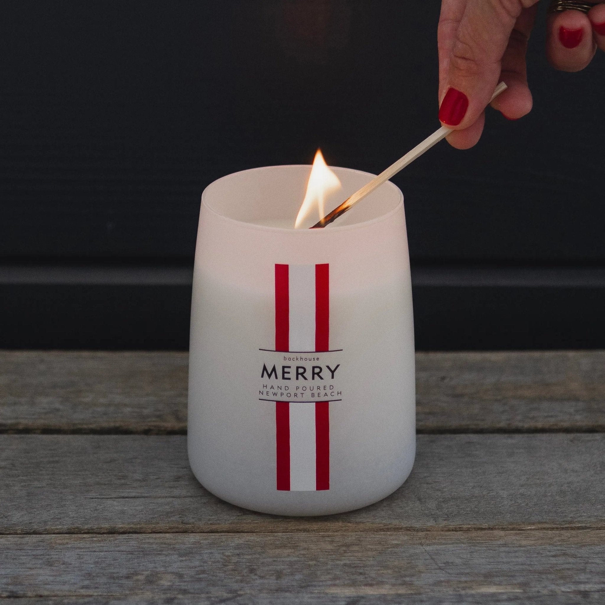 Hand lighting the Merry Mr. Douglas pine candle by backhouse against a dark background.