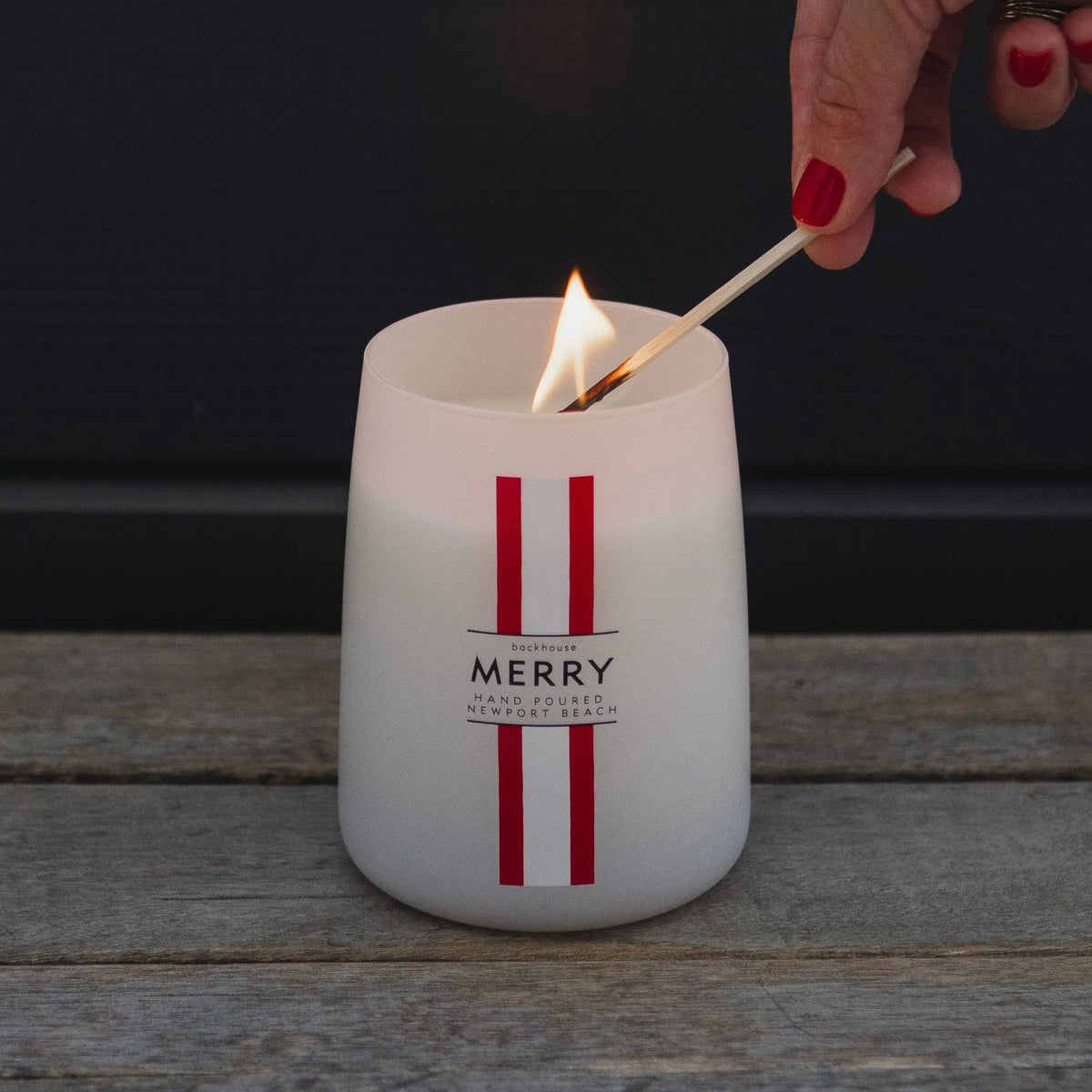 Hand lighting the Merry Mr. Douglas pine candle by backhouse against a dark background.